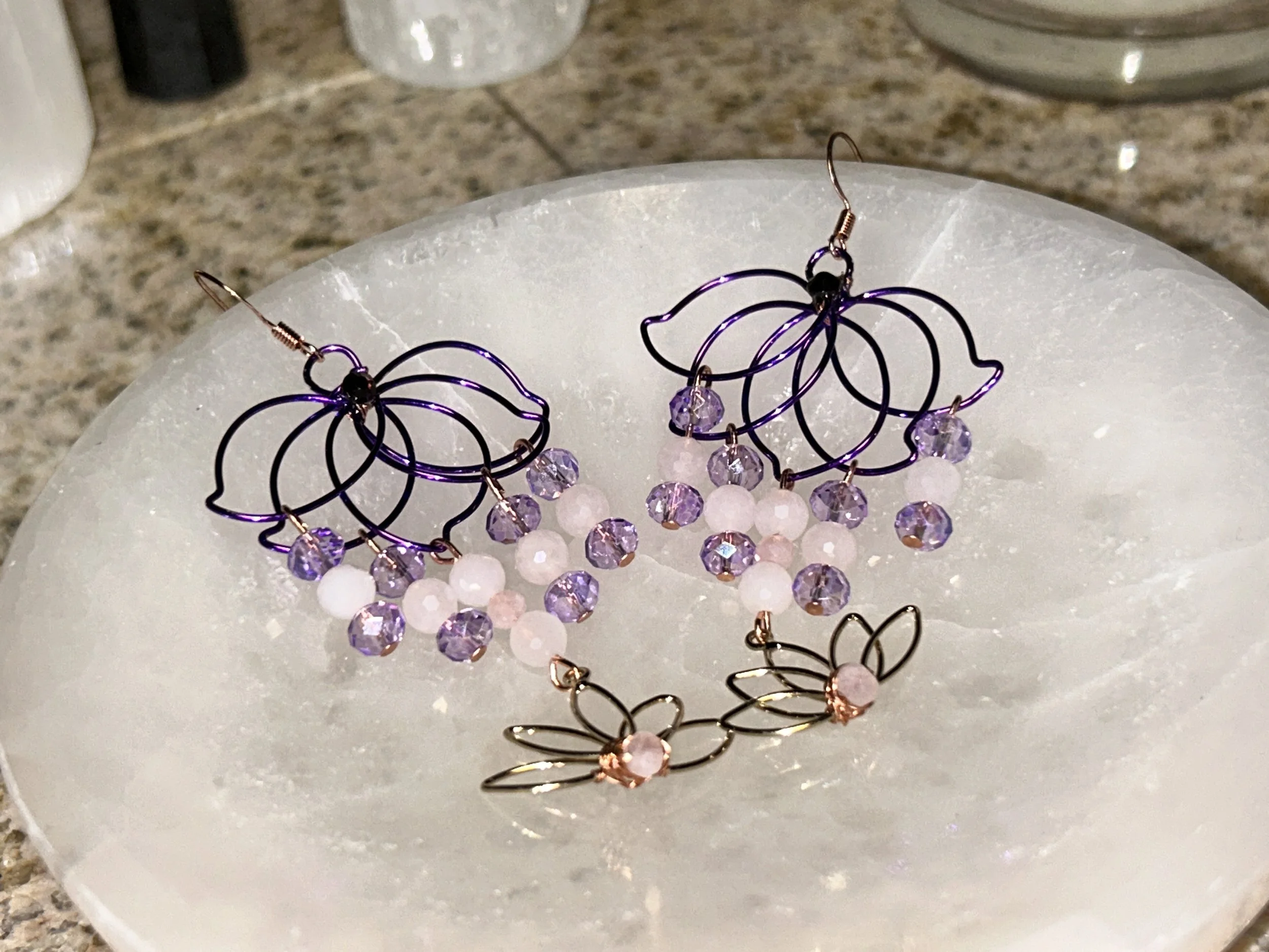 A pair of decorative earrings with purple wire, pink beads, and purple crystal beads, placed on a white circular surface.