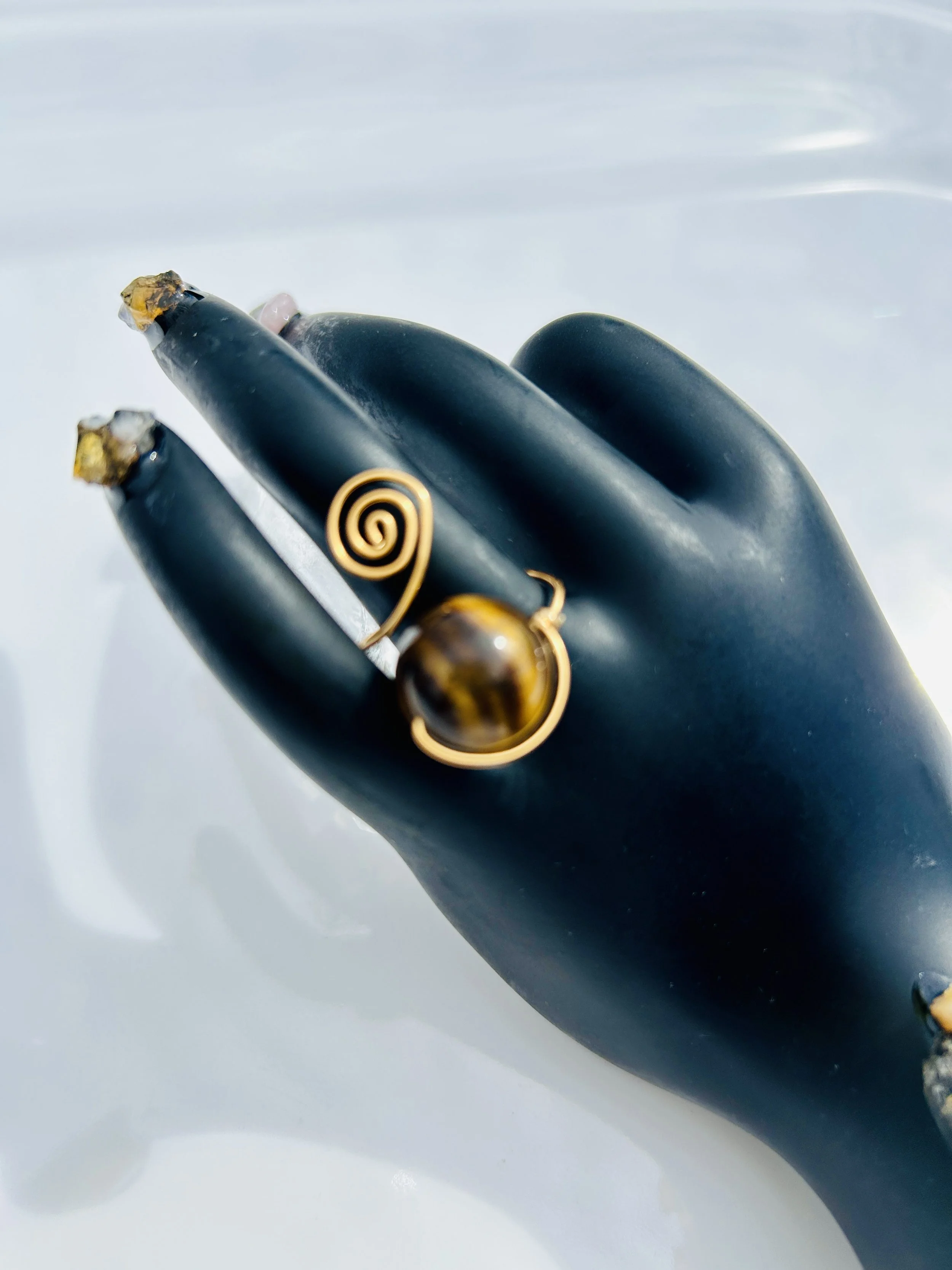 A black mannequin hand displaying a gold wire spiral dangle earring with a large round tiger's eye gemstone.
