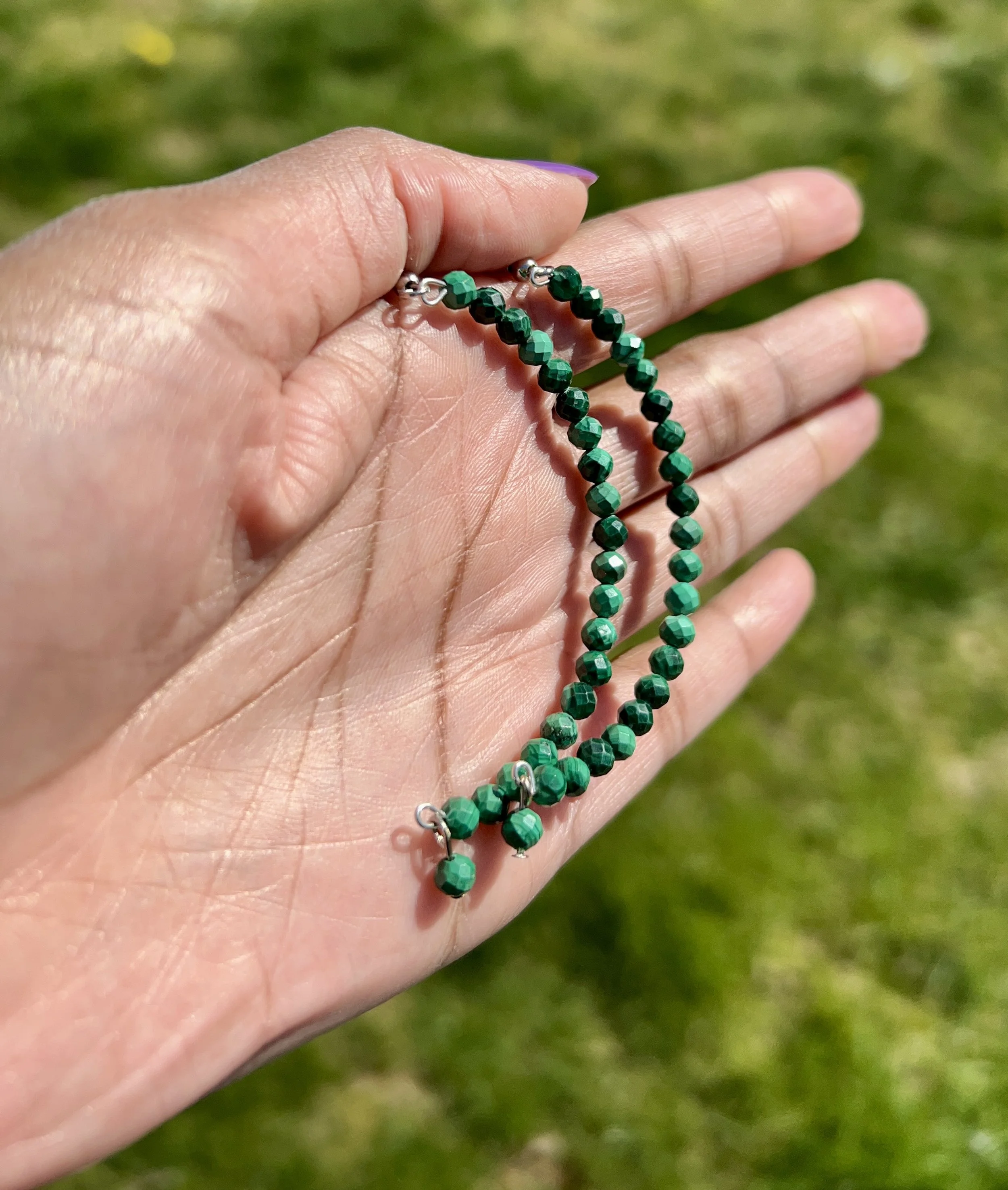 Hand holding a green beaded necklace against a blurred grassy background.
