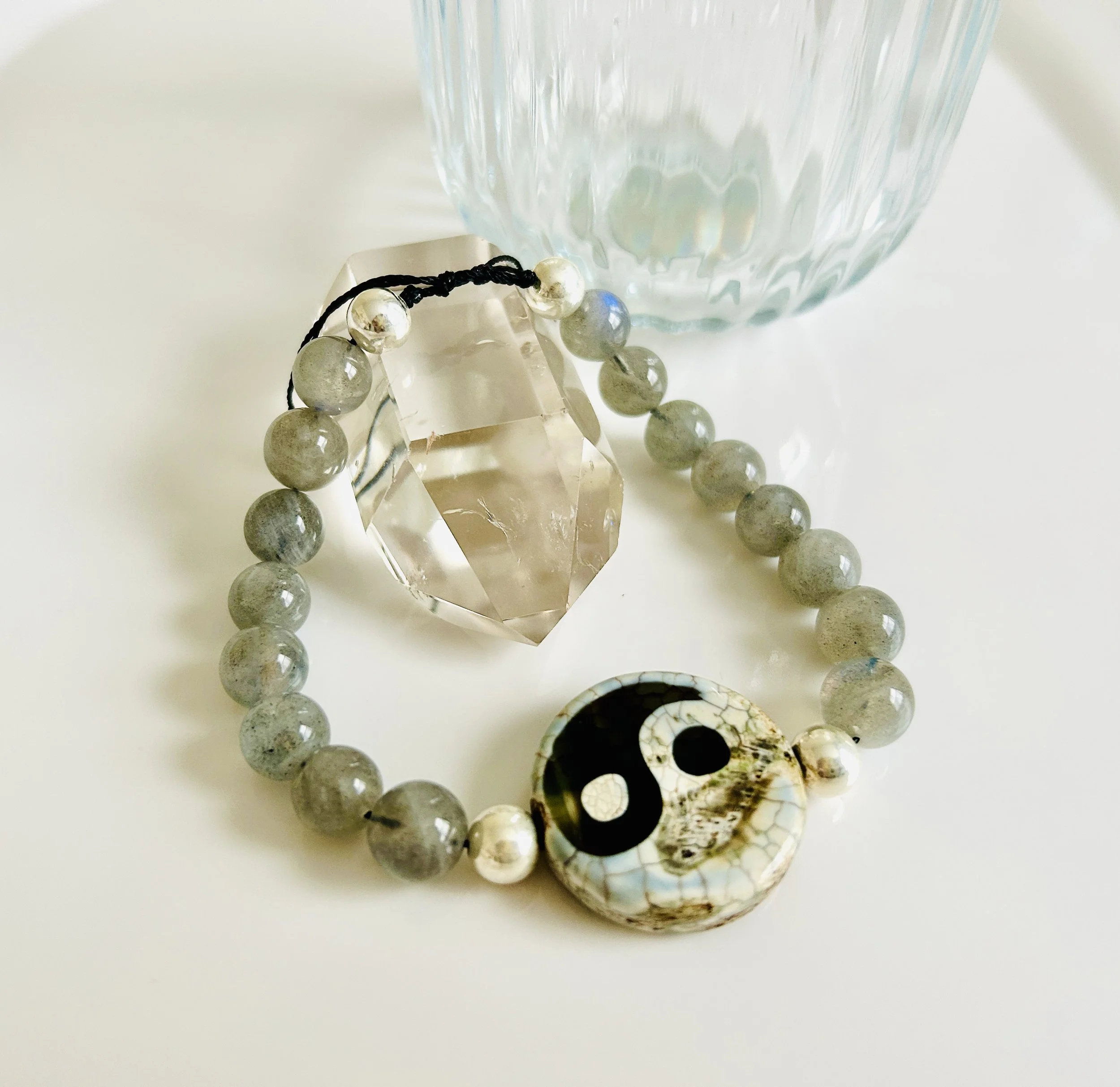 A beaded bracelet with a yin-yang pendant, a large faceted clear crystal, and small silver beads, next to a blue-glass textured cup on a white surface.