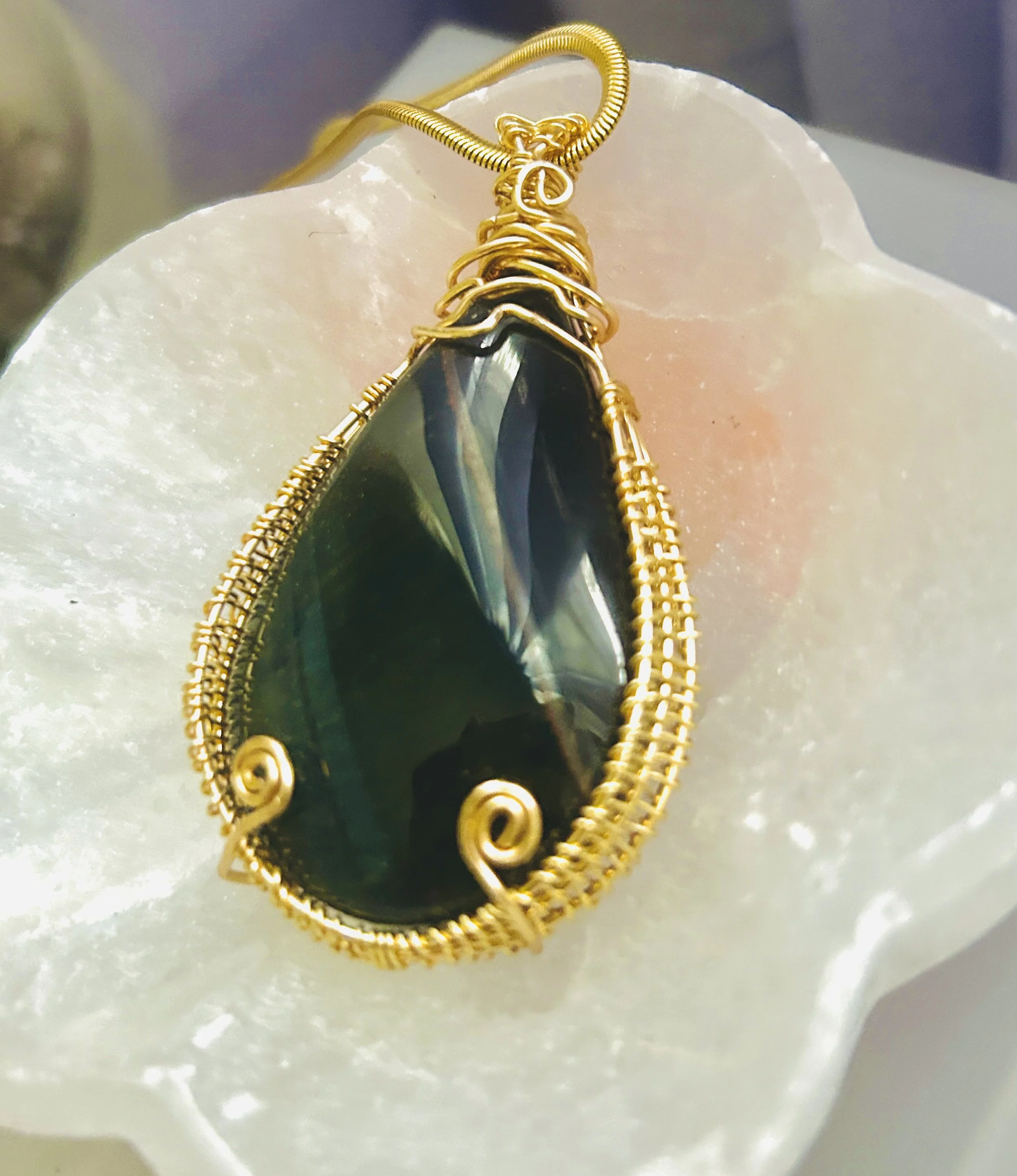 A gold wire-wrapped pendant featuring a large, dark green, teardrop-shaped gemstone, resting on a white flower petal.
