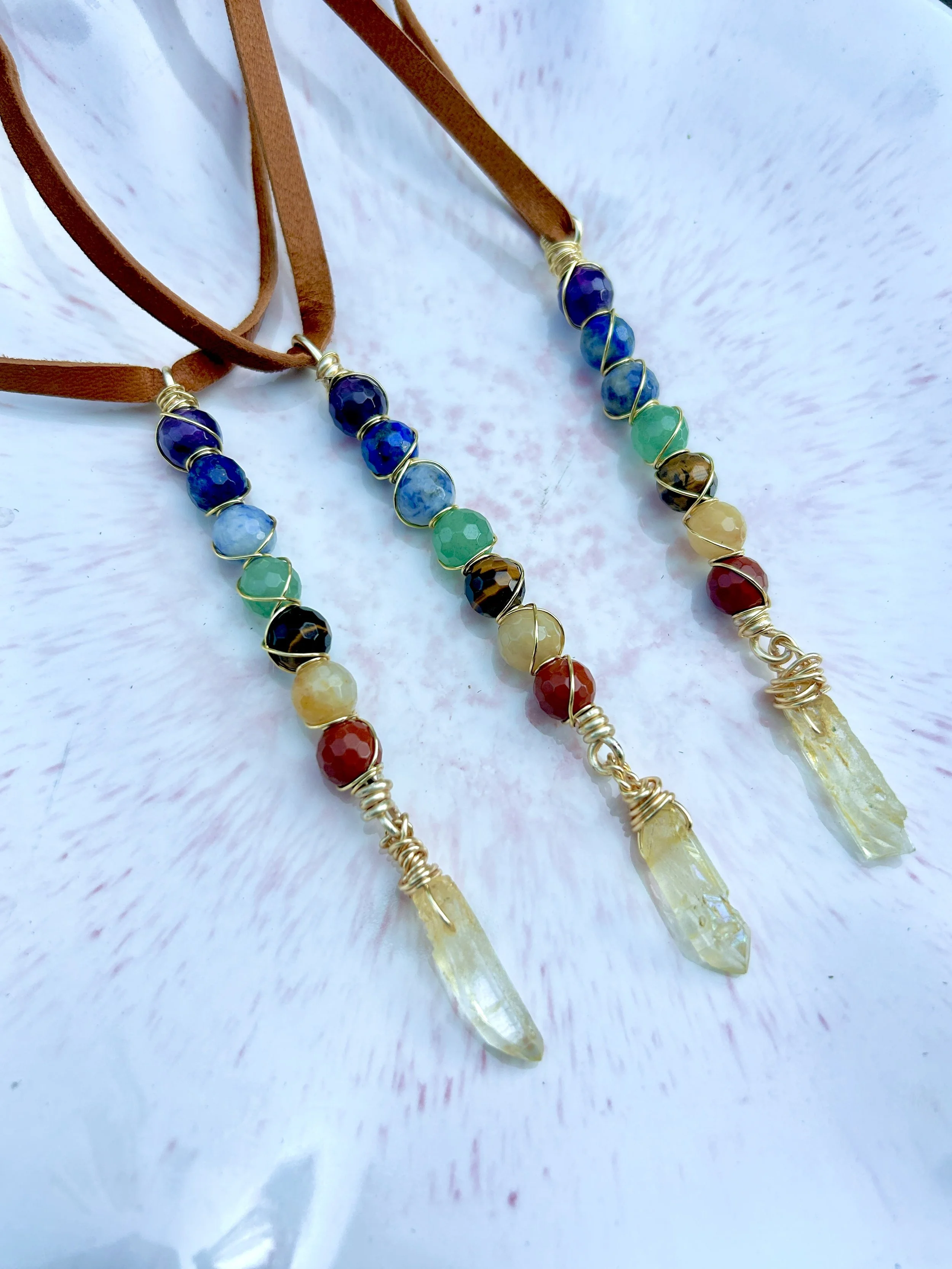 Two rainbow-colored beaded necklaces with leather straps, featuring wire-wrapped beads and quartz crystal pendants, on a white textured surface.