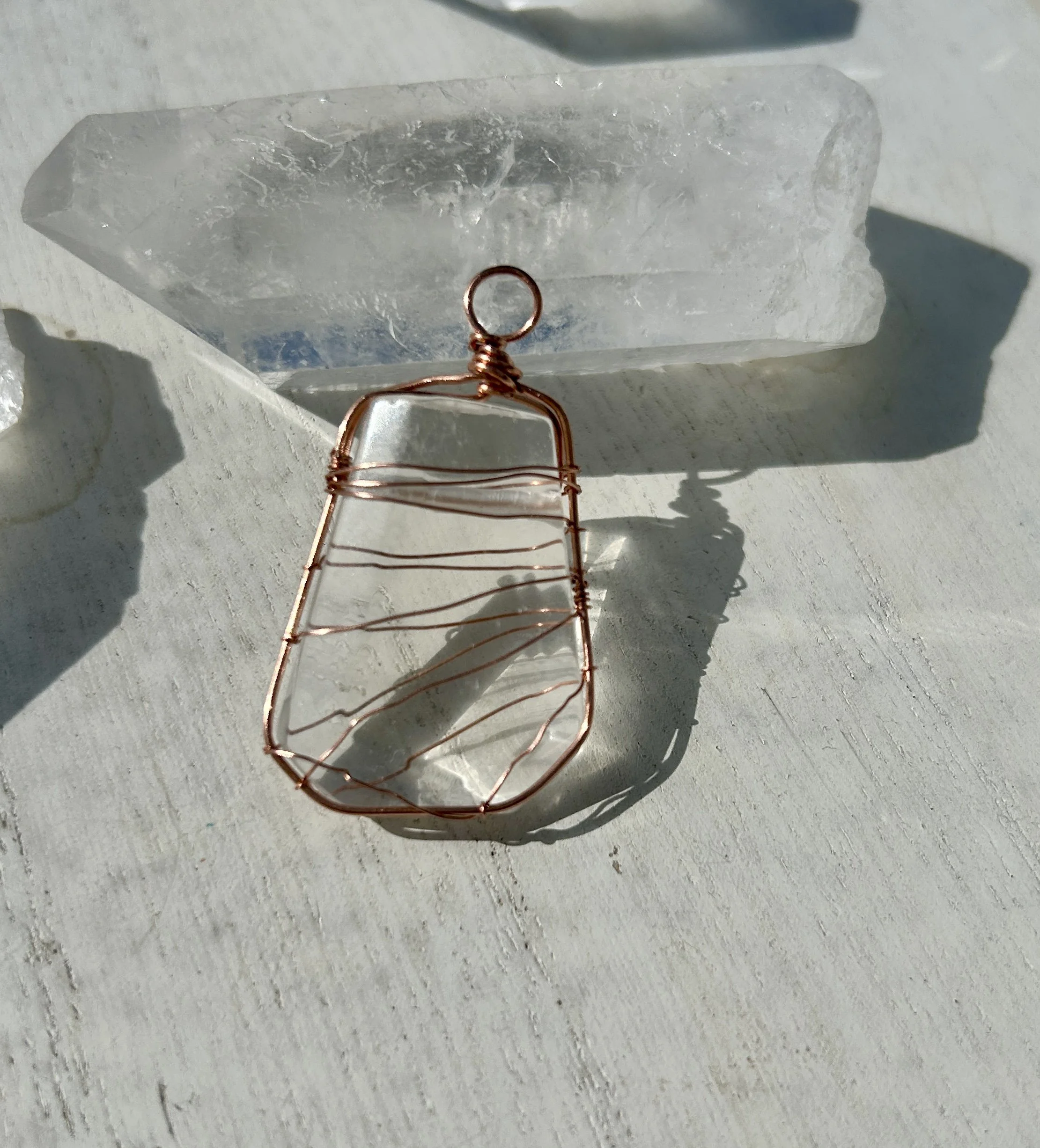 A wire-wrapped pendant with a clear, rectangular stone, resting on a white surface, placed next to a large clear quartz crystal.