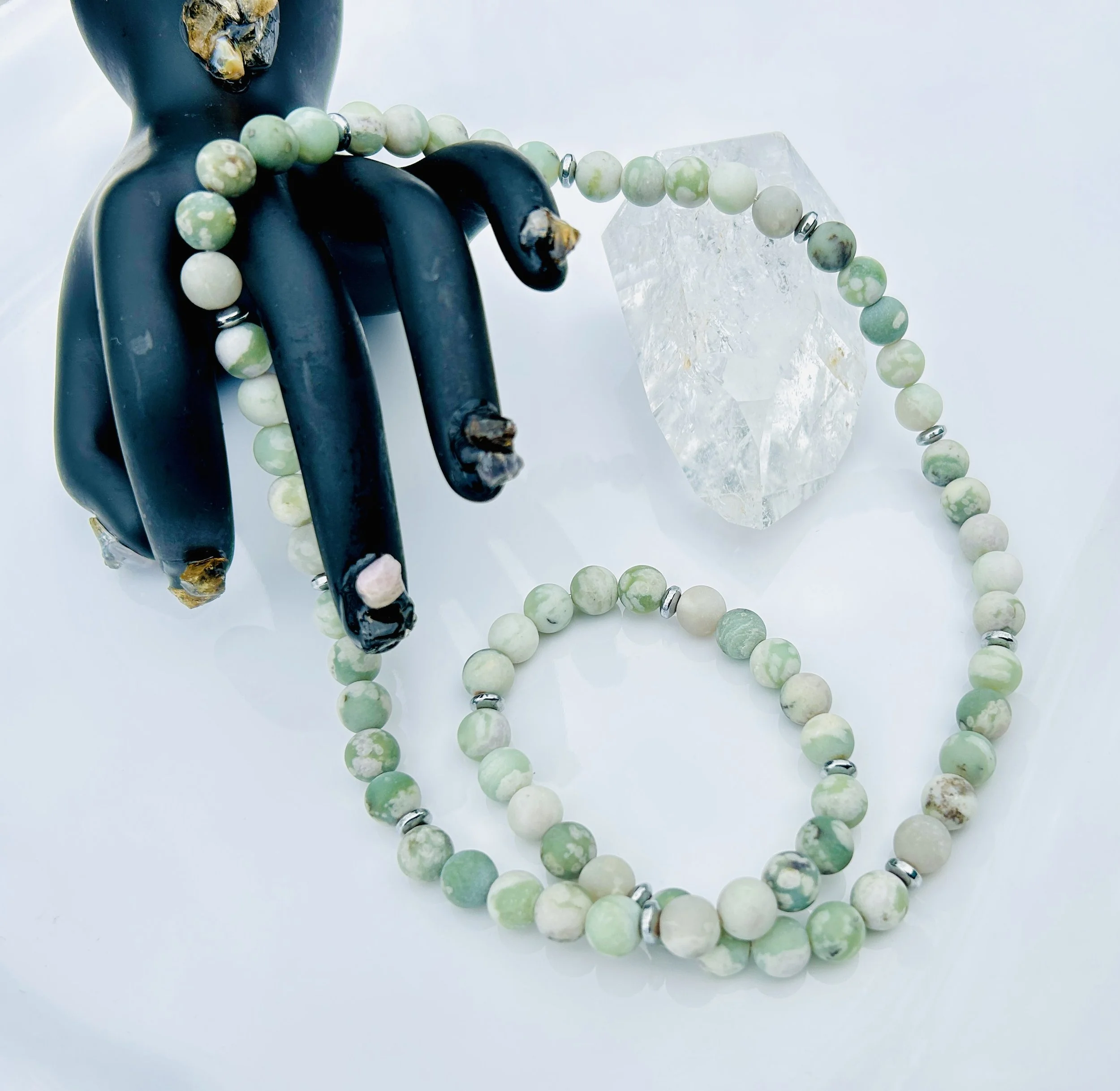 A black claw-shaped jewelry holder with a green and white beaded necklace draped over it, a clear quartz crystal, and a white background.