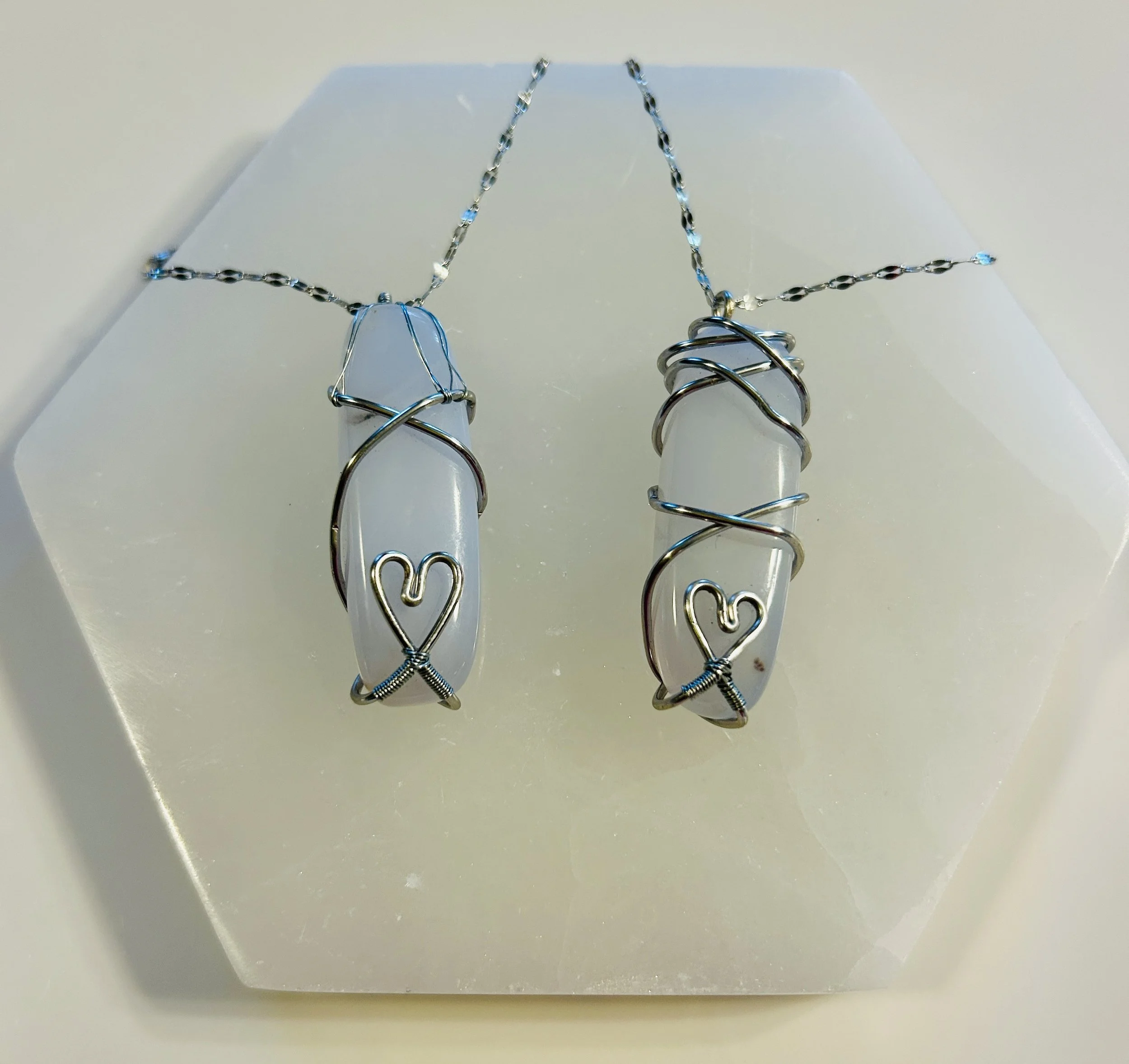 Two jewelry pendants with white stones wrapped in silver wire, each featuring a heart-shaped design at the bottom, on a chain displayed on a white surface.