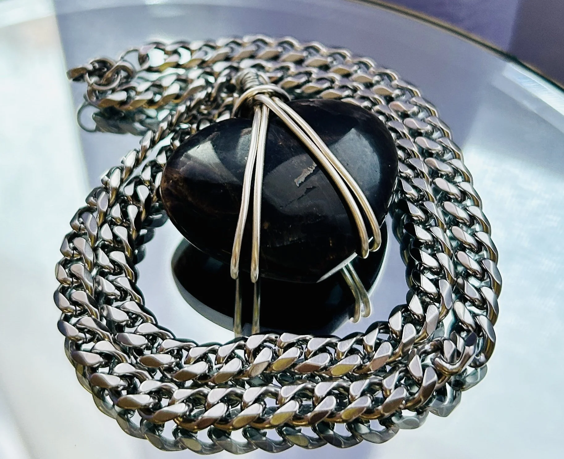 A black polished stone wrapped with silver wire, resting on a smaller black stone, surrounded by a thick silver chain, on a reflective glass surface.