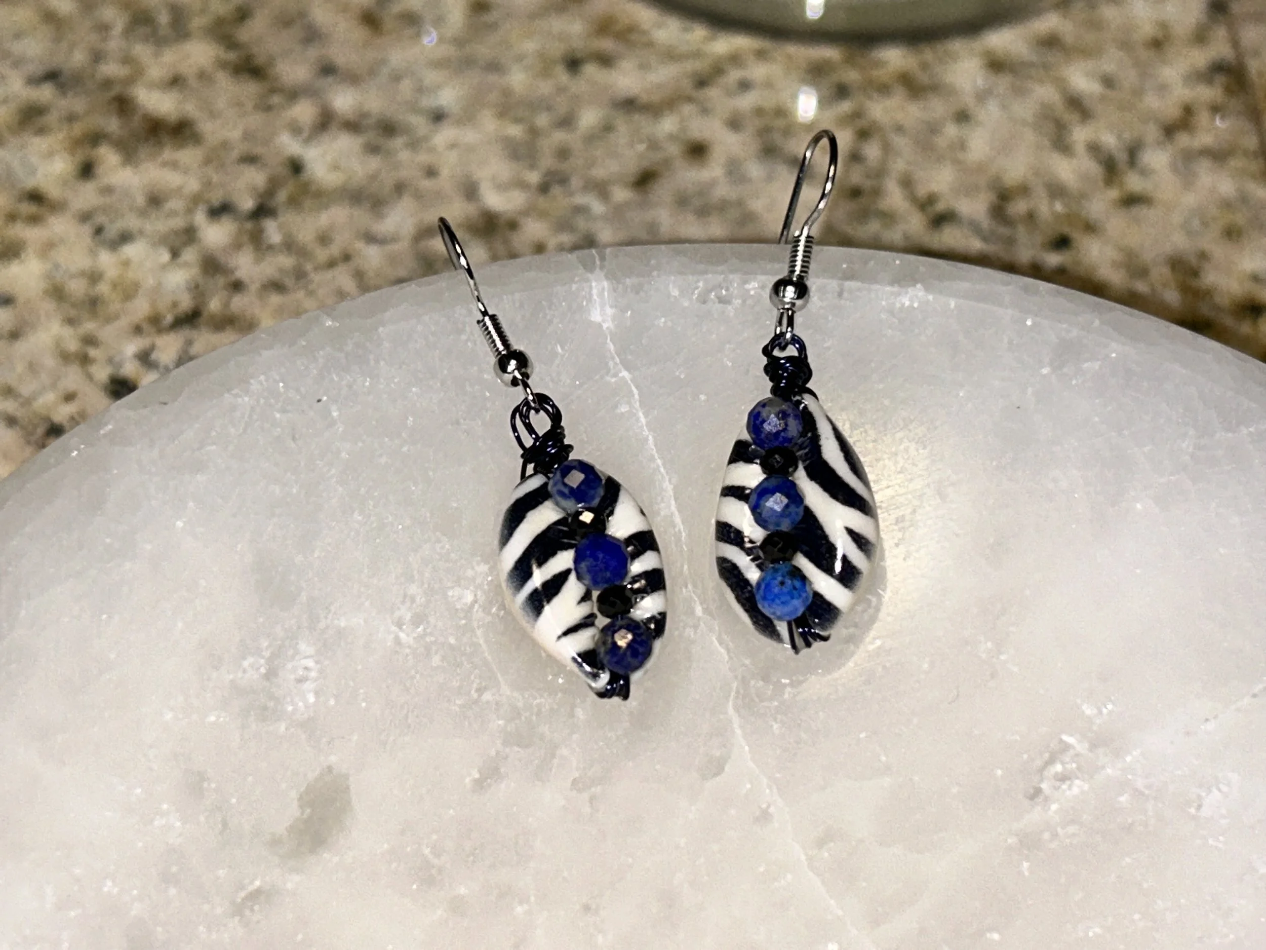 Pair of earrings with black and white zebra stripe design and small blue beads, displayed on a white, textured surface.