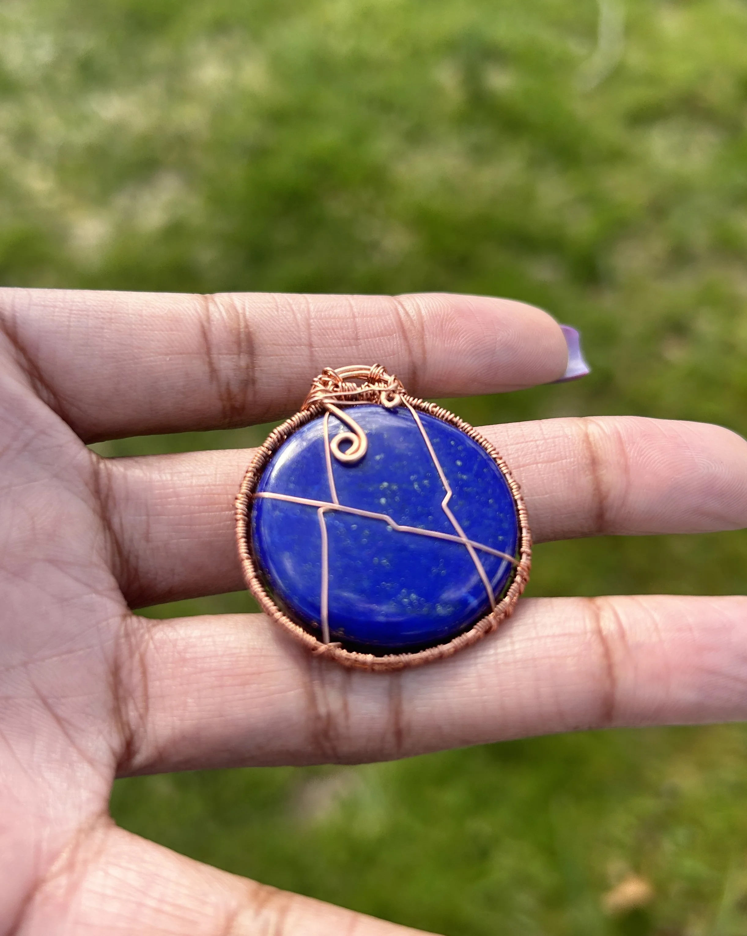 A person holding a round blue gemstone necklace wrapped in copper wire, with an outdoor grassy background.