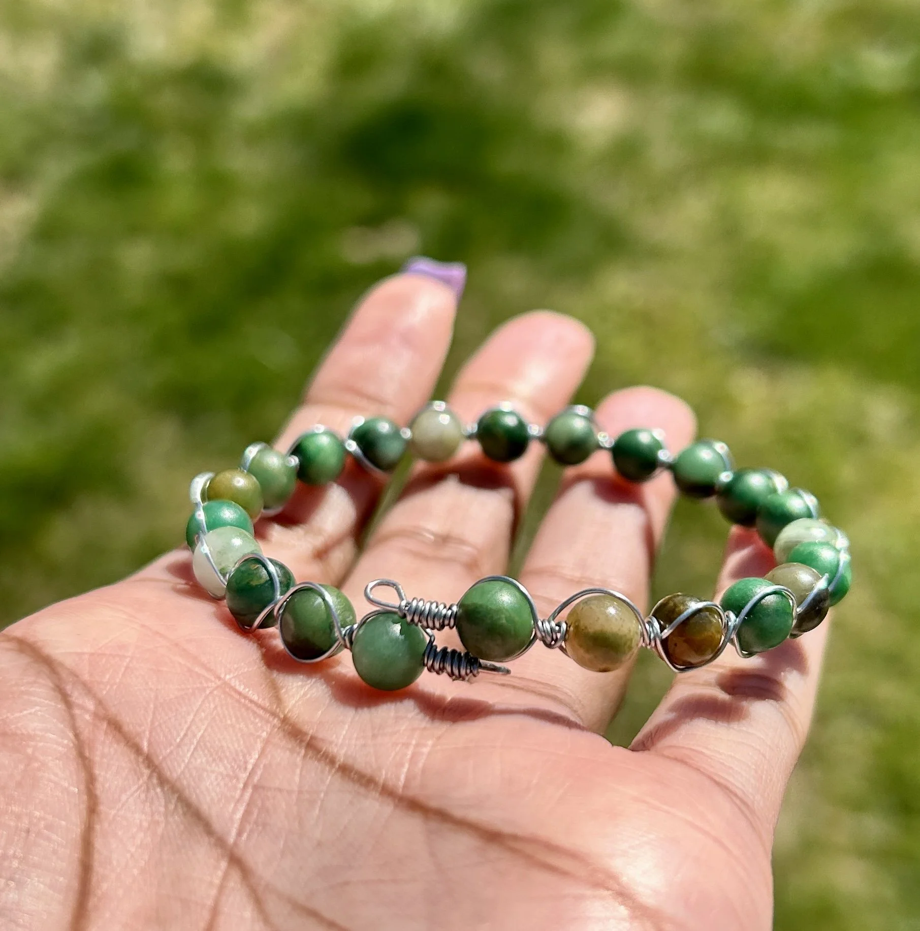 Hand holding a green beaded bracelet outdoors with a grassy background.