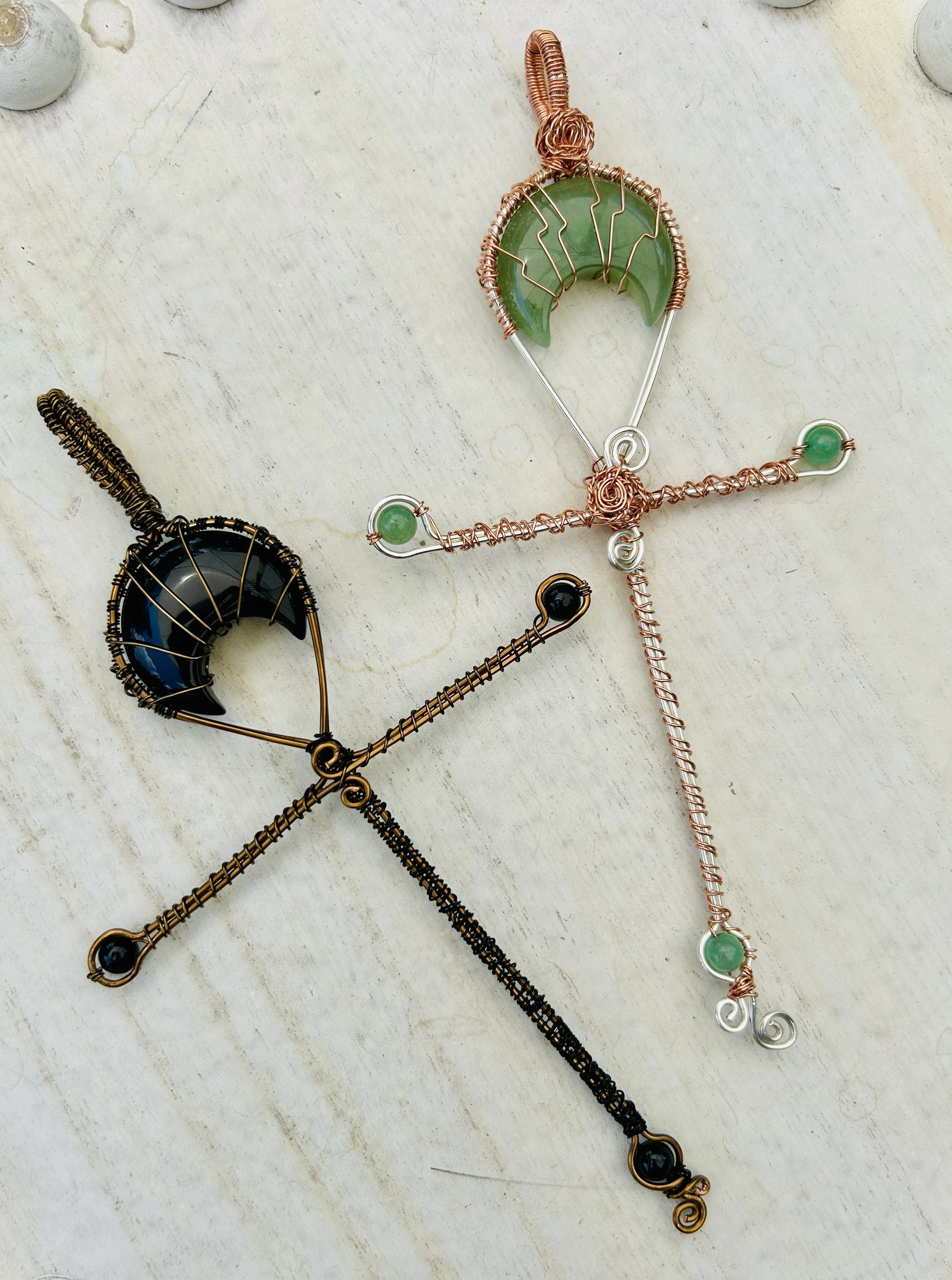 Wire-wrapped decorative angel ornaments with crescent moon shapes, made with copper and black or green stones, placed on a light-colored wooden surface.