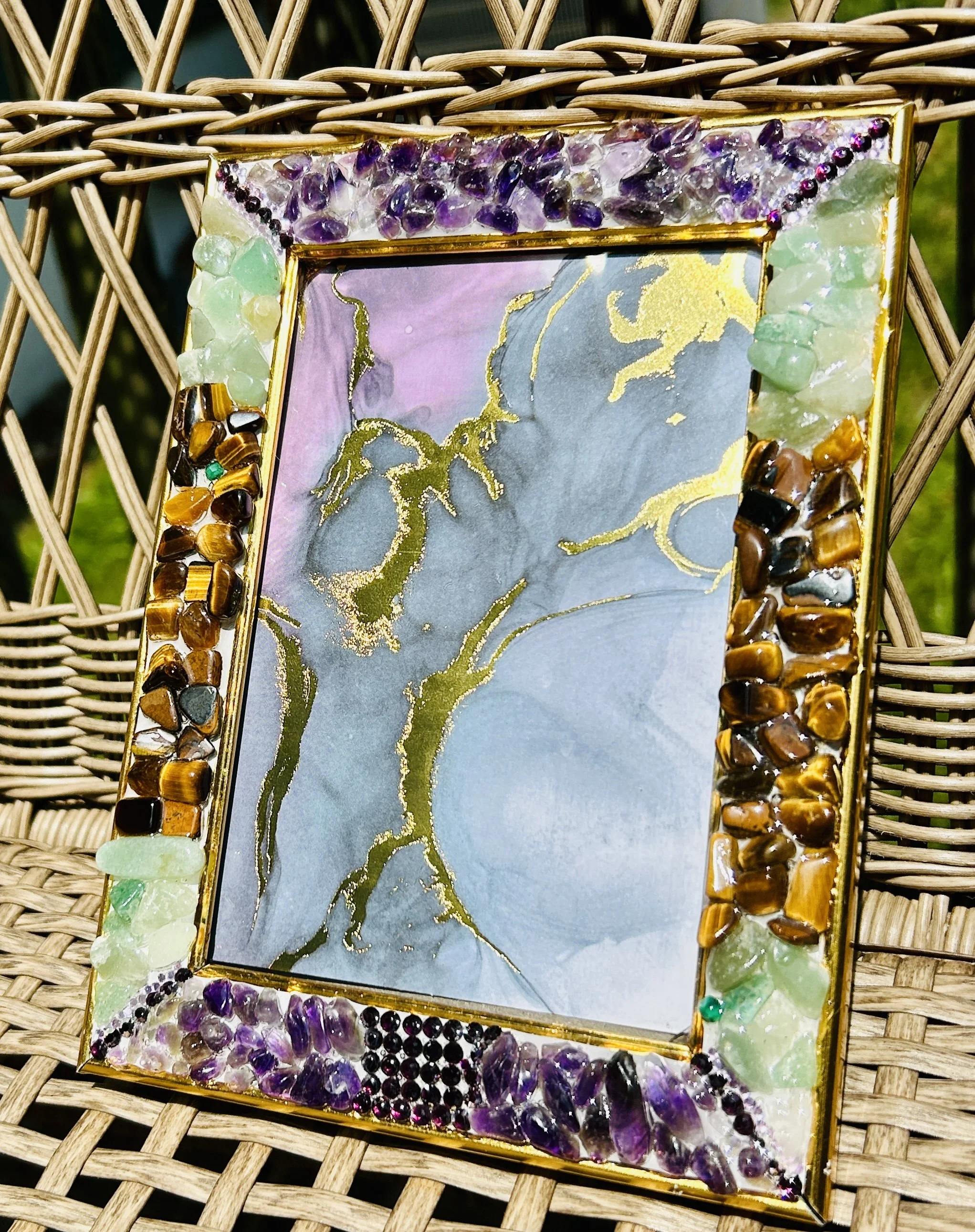 Decorative picture frame adorned with colorful stones and crystals, with a marbled abstract background inside it. The frame is set on a wicker surface.