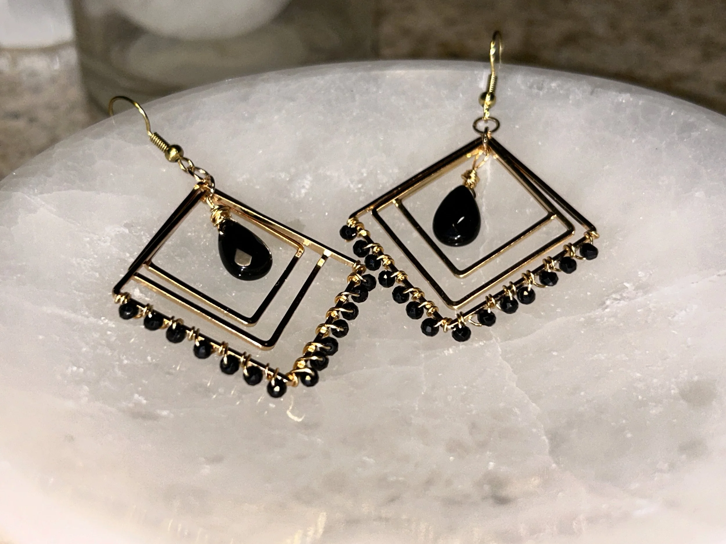 Gold geometric earrings with black teardrop pendants and small black beads hanging from the edges, placed on a white surface.