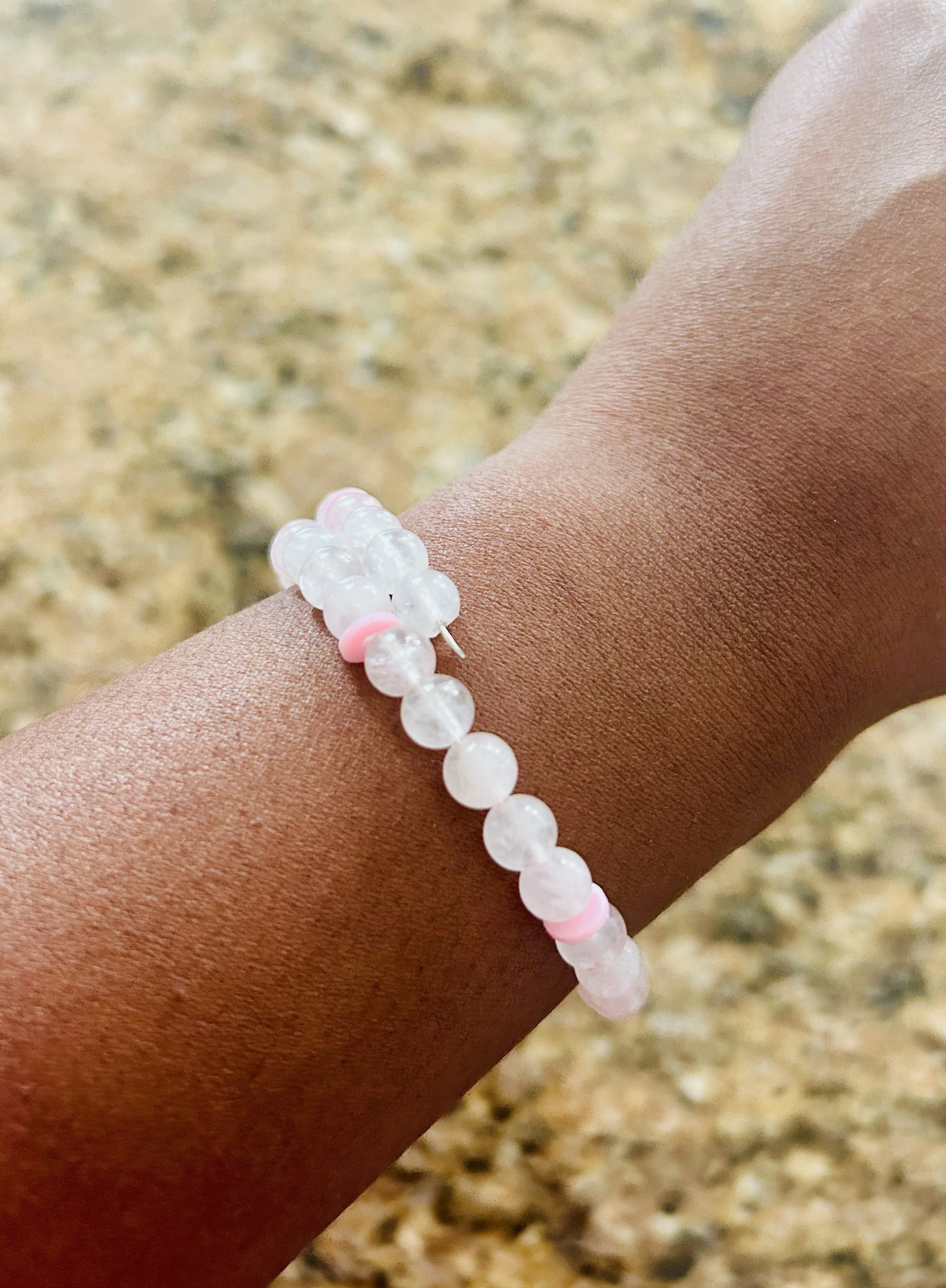 Close-up of a tan arm wearing a white beaded bracelet with pink accents, against a blurred outdoor background of sandy or rocky ground.