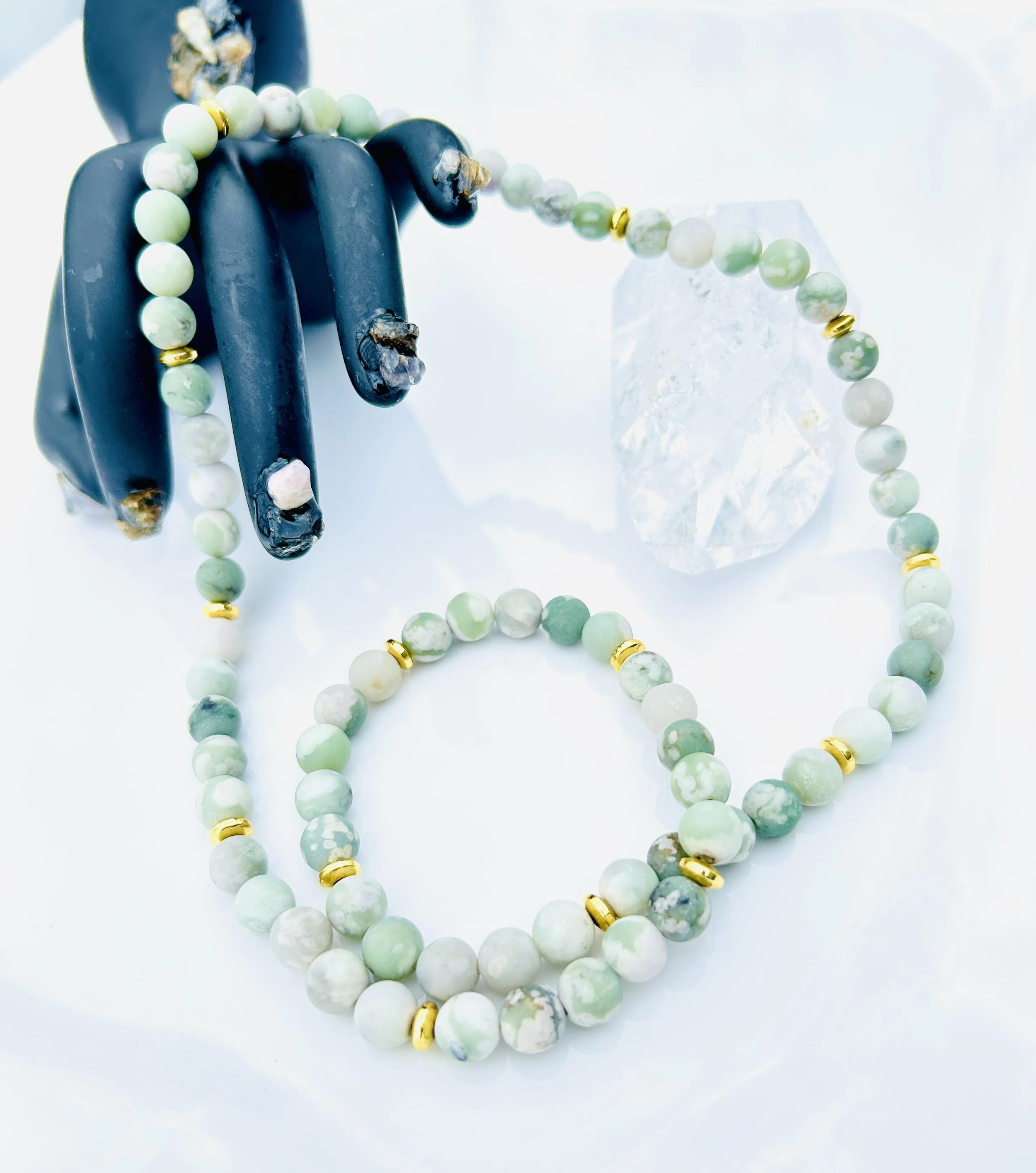 A black mannequin hand with navy blue painted nails, wearing a green and white beaded necklace with gold spacers, alongside a matching bracelet, and a large clear quartz crystal on a white surface.