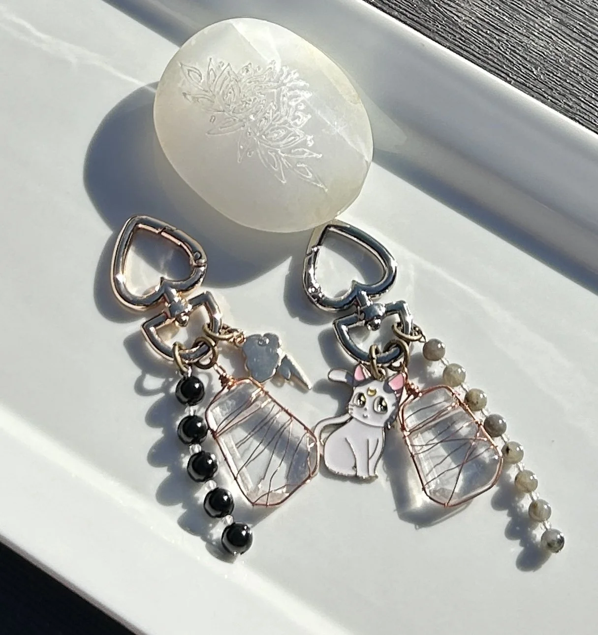 A pair of heart-shaped keychains with various charms, including a cat, a leaf, beads, and wire-wrapped stones, on a white surface with a decorative soap or stone and a window.
