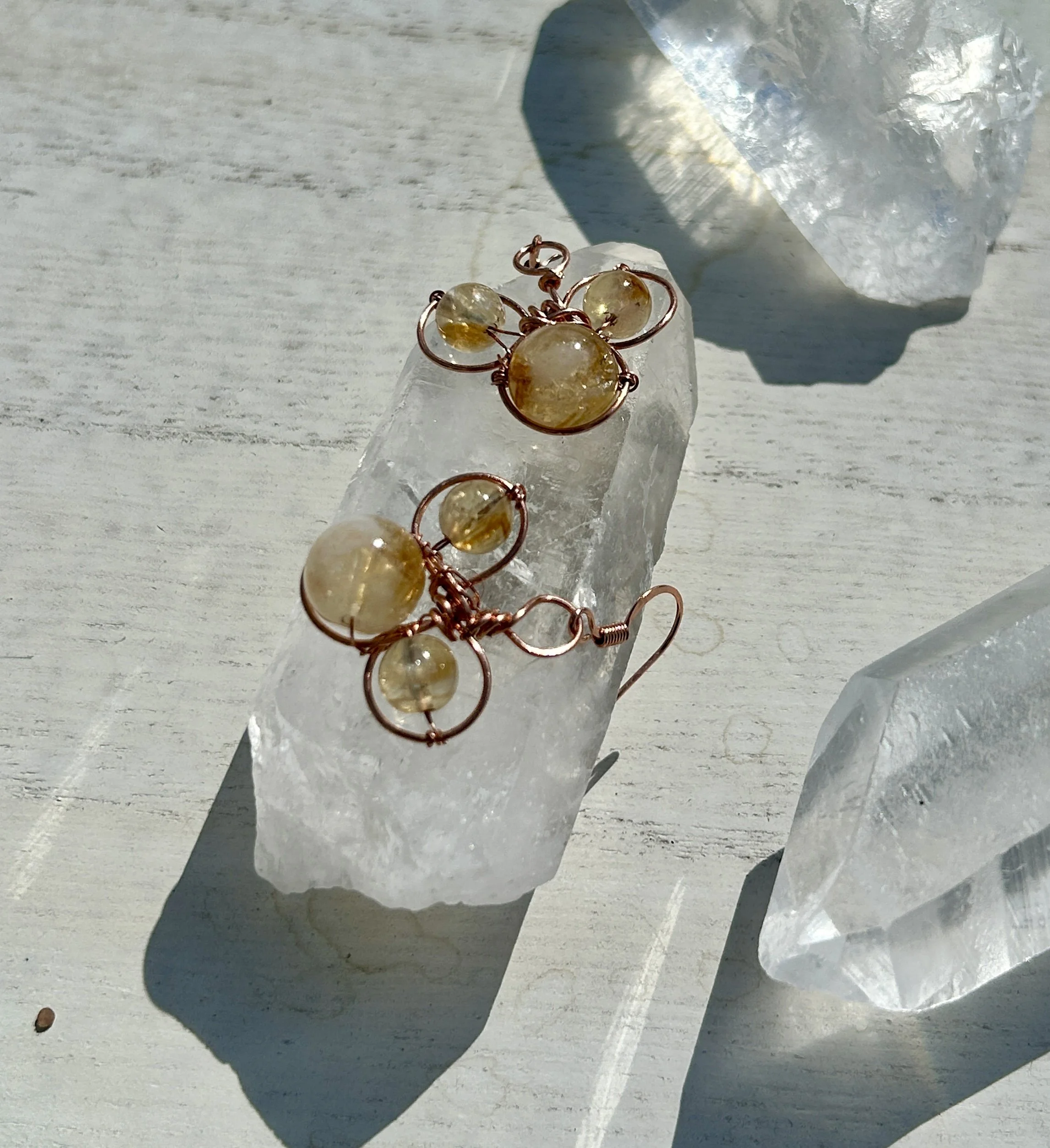 Gold and clear gemstone jewelry, including earrings and a pendant, resting on a large clear quartz crystal with sunlight casting shadows.