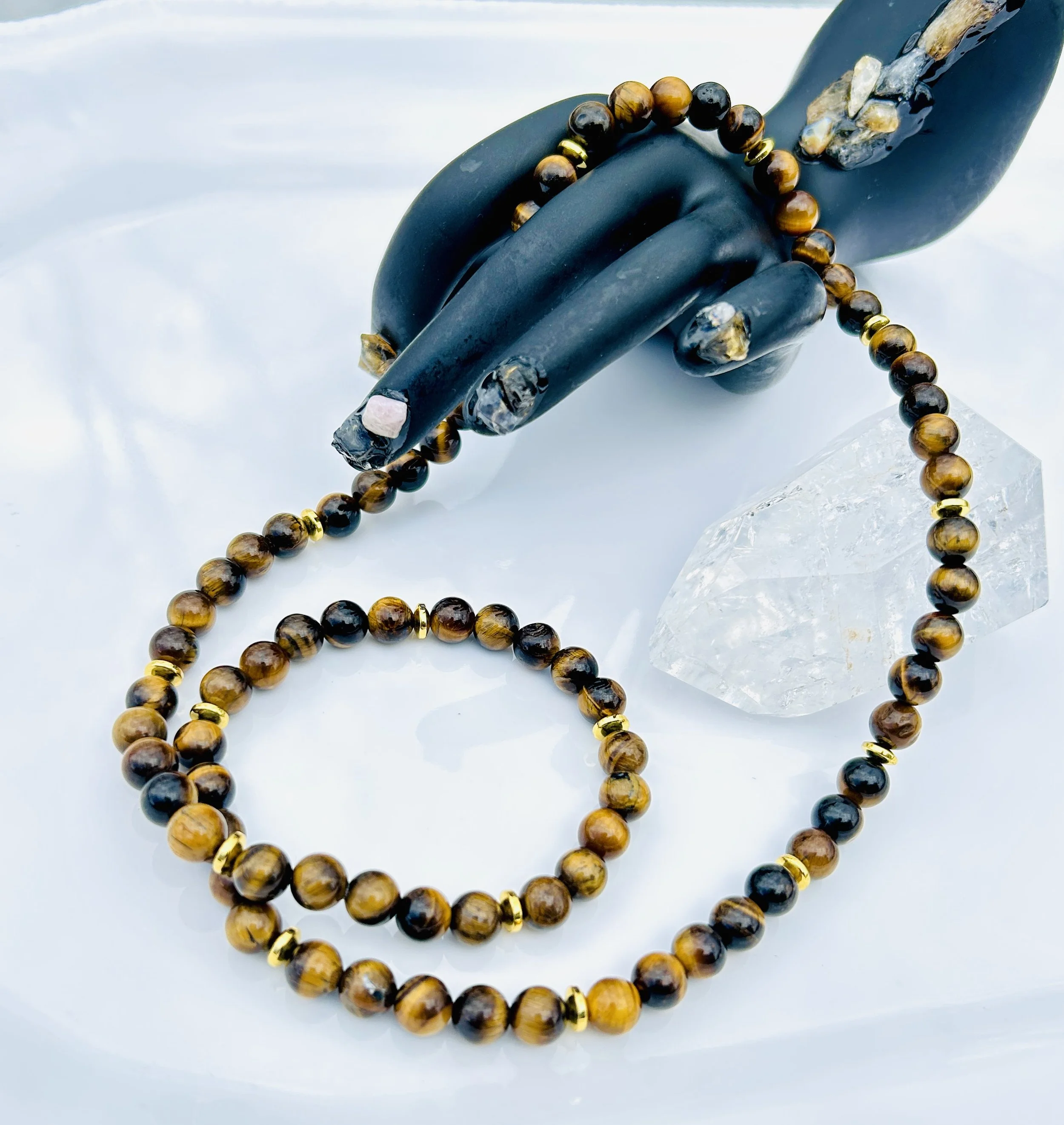 A beaded necklace and bracelet made of tiger's eye stones, displayed on a white surface, with a black hand-shaped jewelry holder and a large clear quartz crystal.