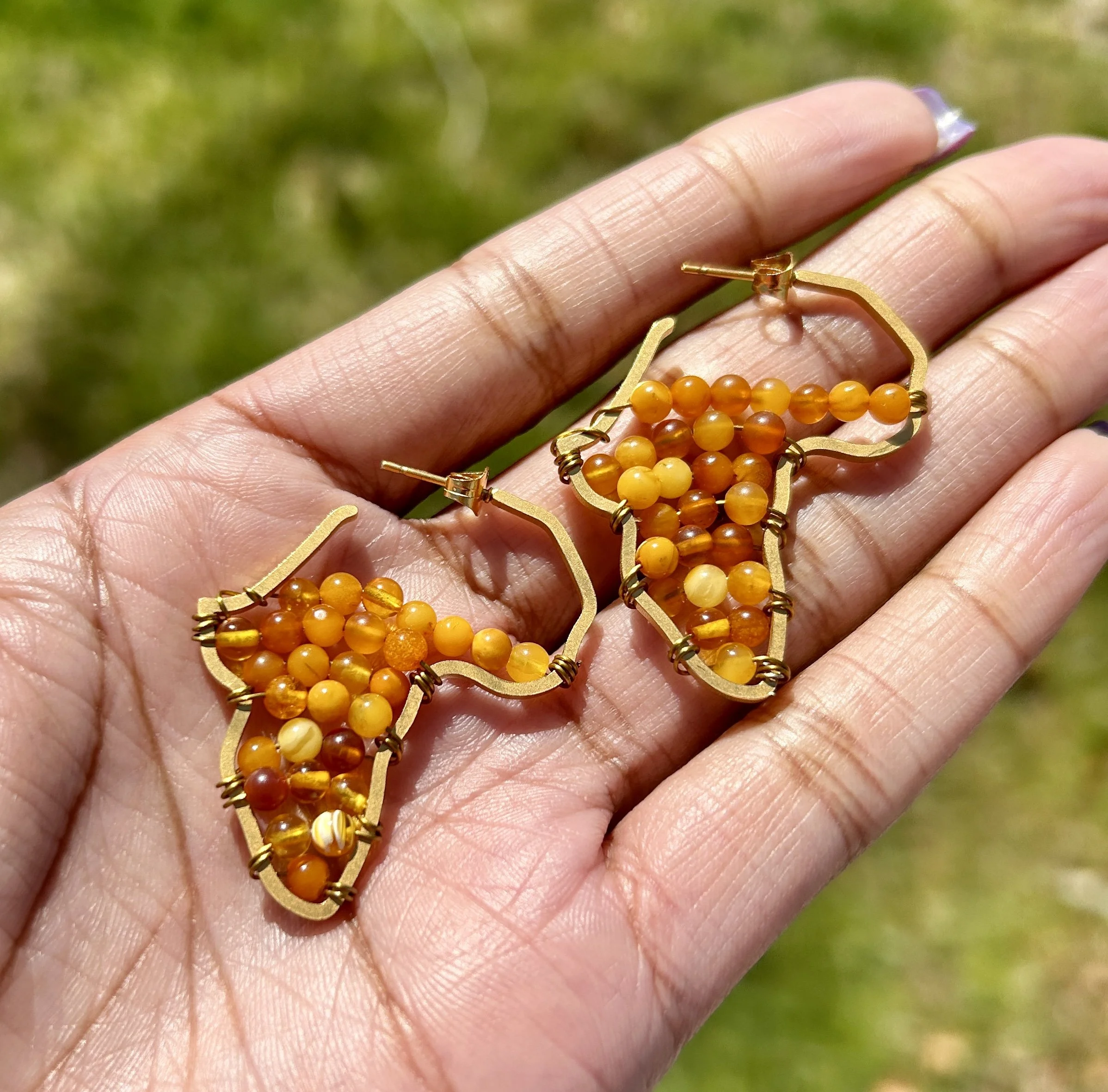 Gold earrings with orange and yellow beads held on a person's hand against a blurred green outdoor background.