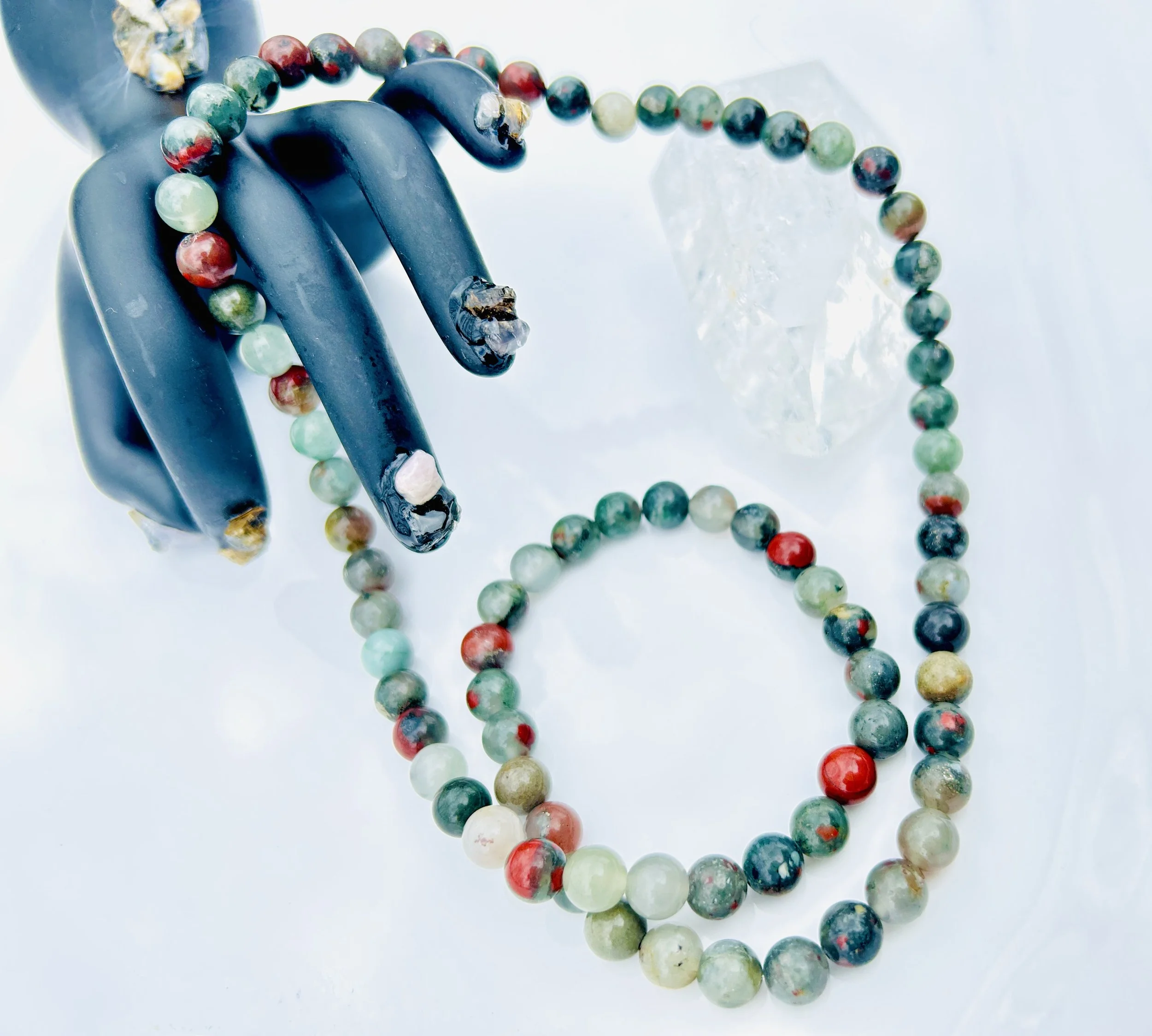 A beaded necklace with multicolored round beads, draped on a black mannequin hand, with a clear quartz crystal nearby.