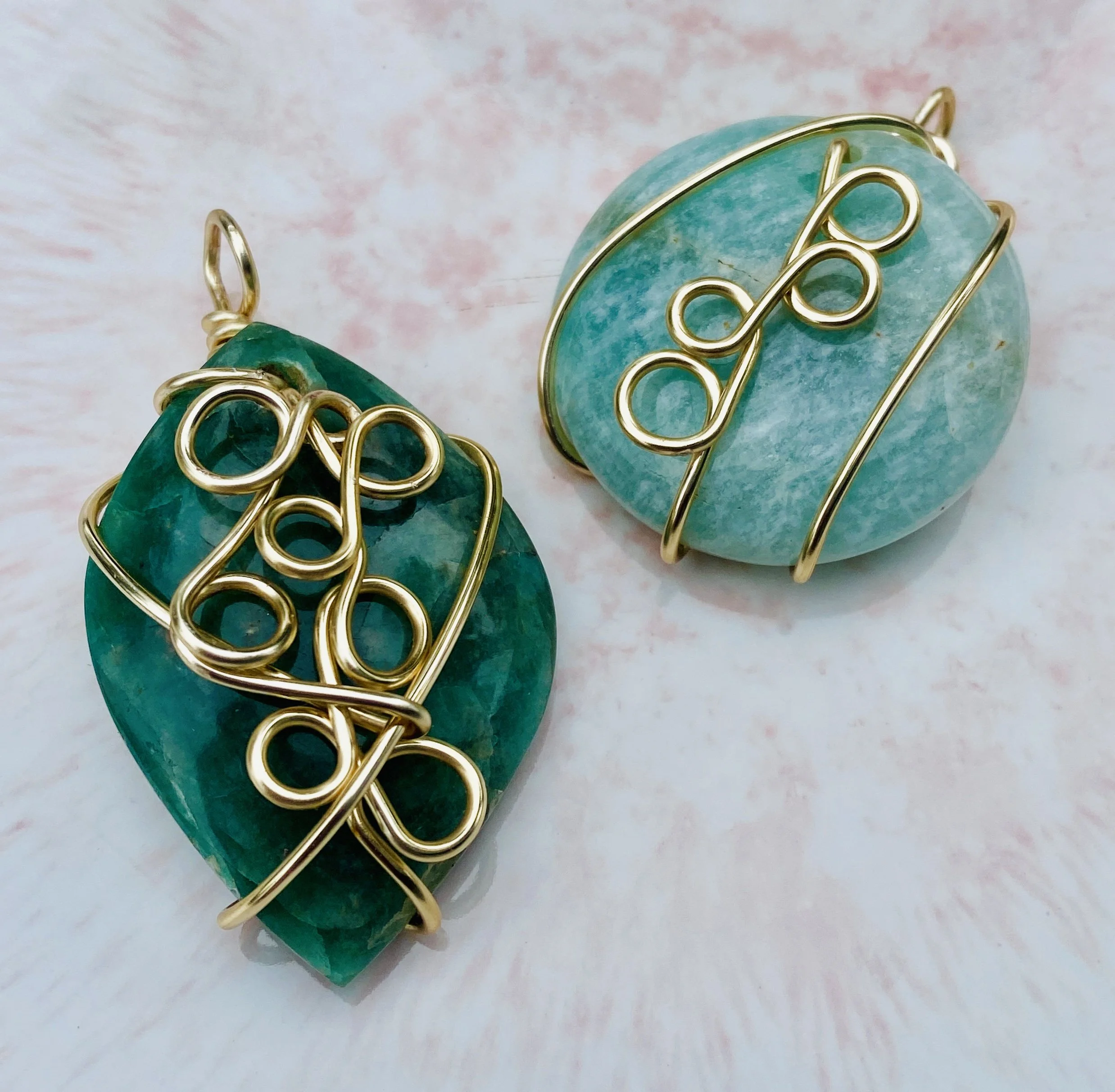 Two turquoise stone pendants wrapped in gold wire on a pinkish background.