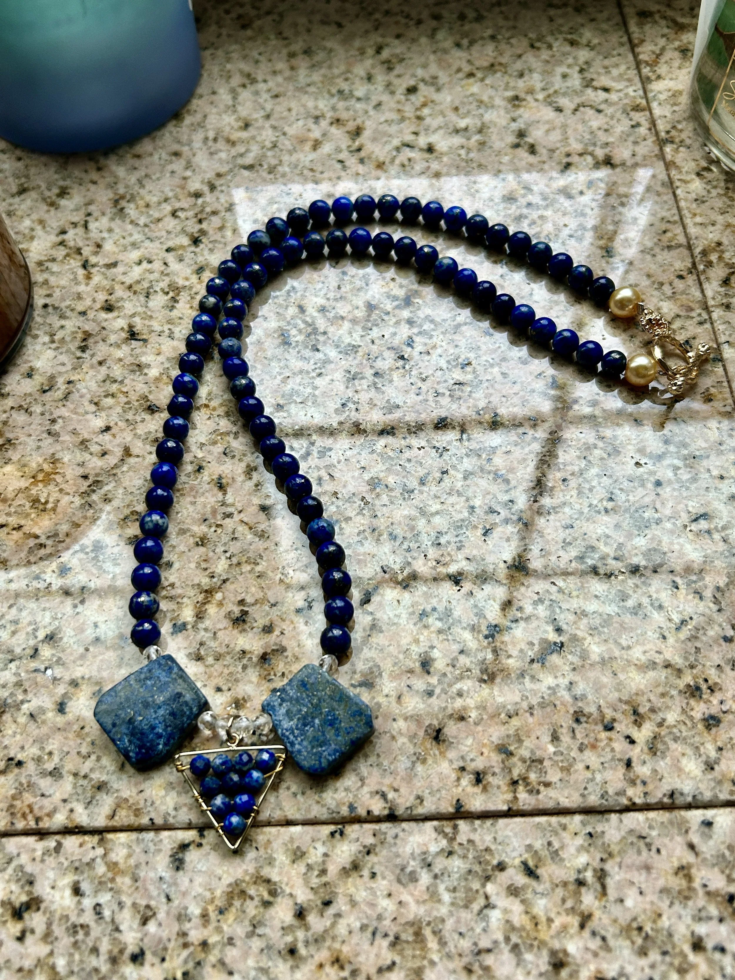 Blue beaded necklace with two large square beads and a triangle pendant with smaller blue beads on a granite surface.