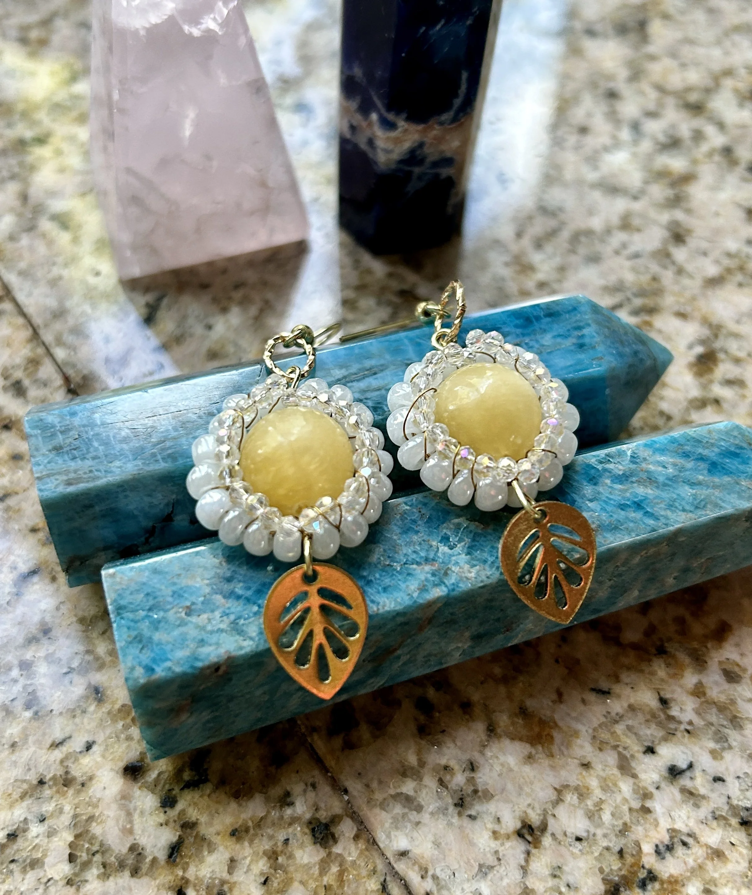 A pair of earrings with yellow stones, surrounded by small white beads and golden leaf charms, placed on a blue textured surface with three crystals in the background on a granite countertop.