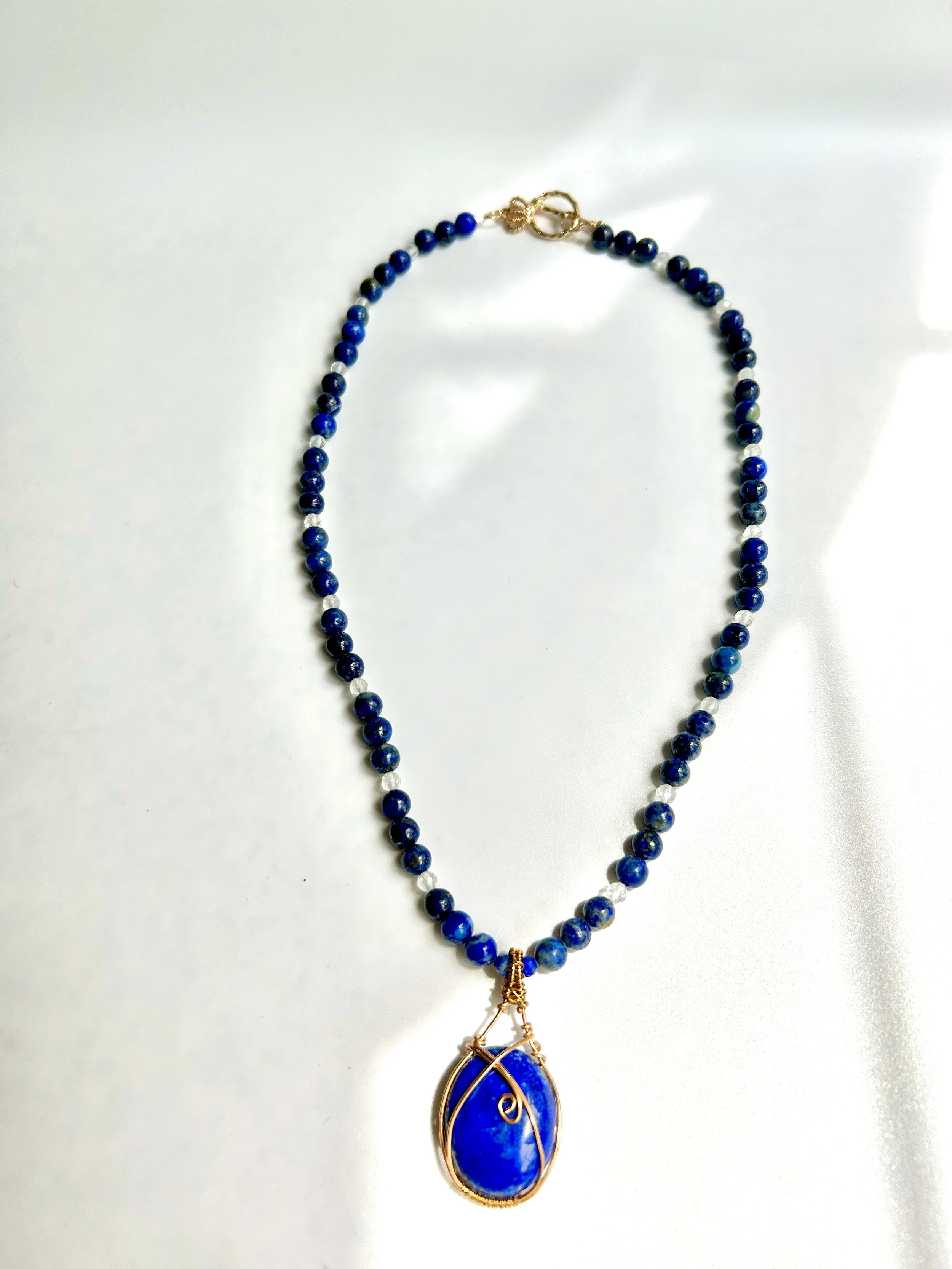 A necklace with blue beads and a large blue pendant with gold wire wrapping on a white background.