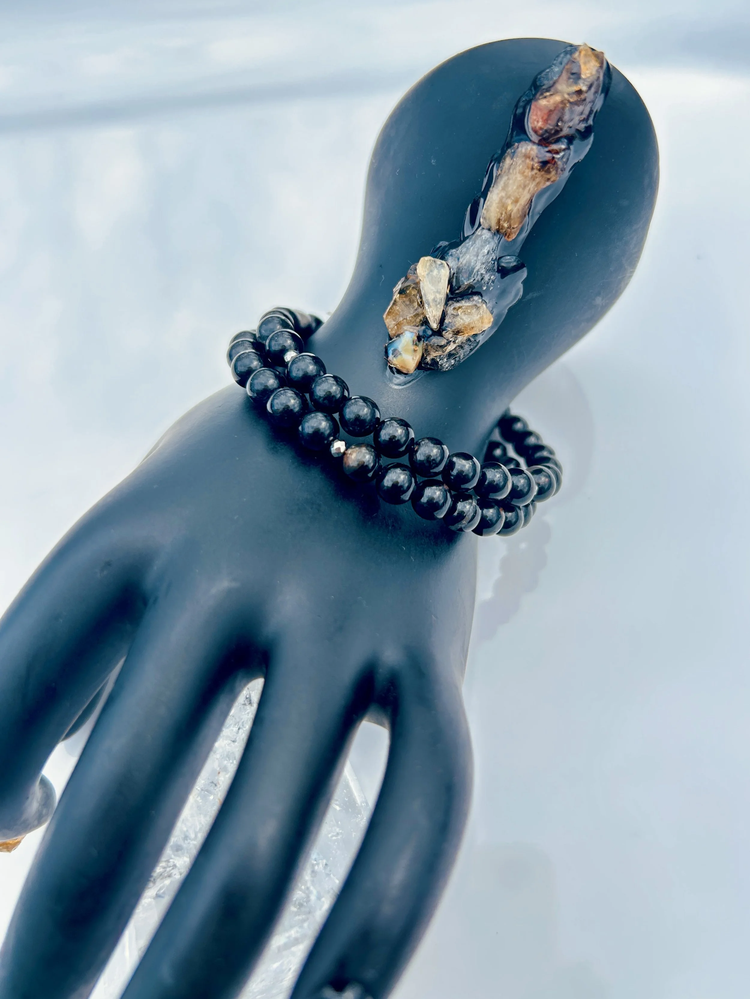 A black mannequin hand wearing a black beaded necklace with a large, irregularly shaped crystal pendant, displayed against a white background.