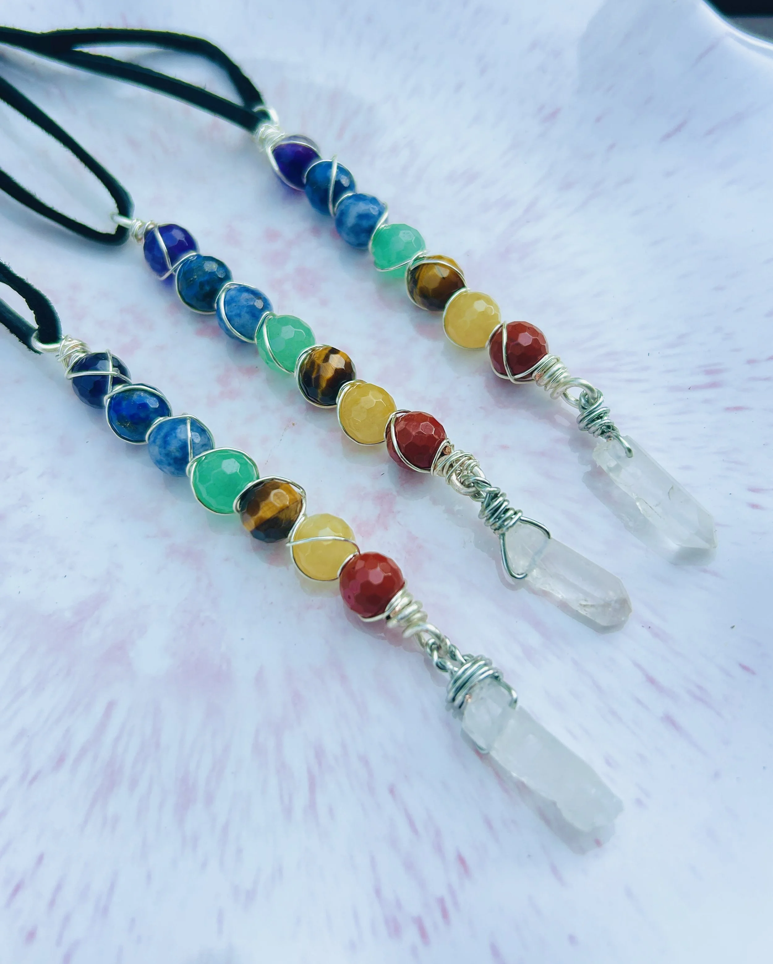 Rainbow-colored beaded necklaces with pendants featuring clear quartz crystals on a light pink and white background.