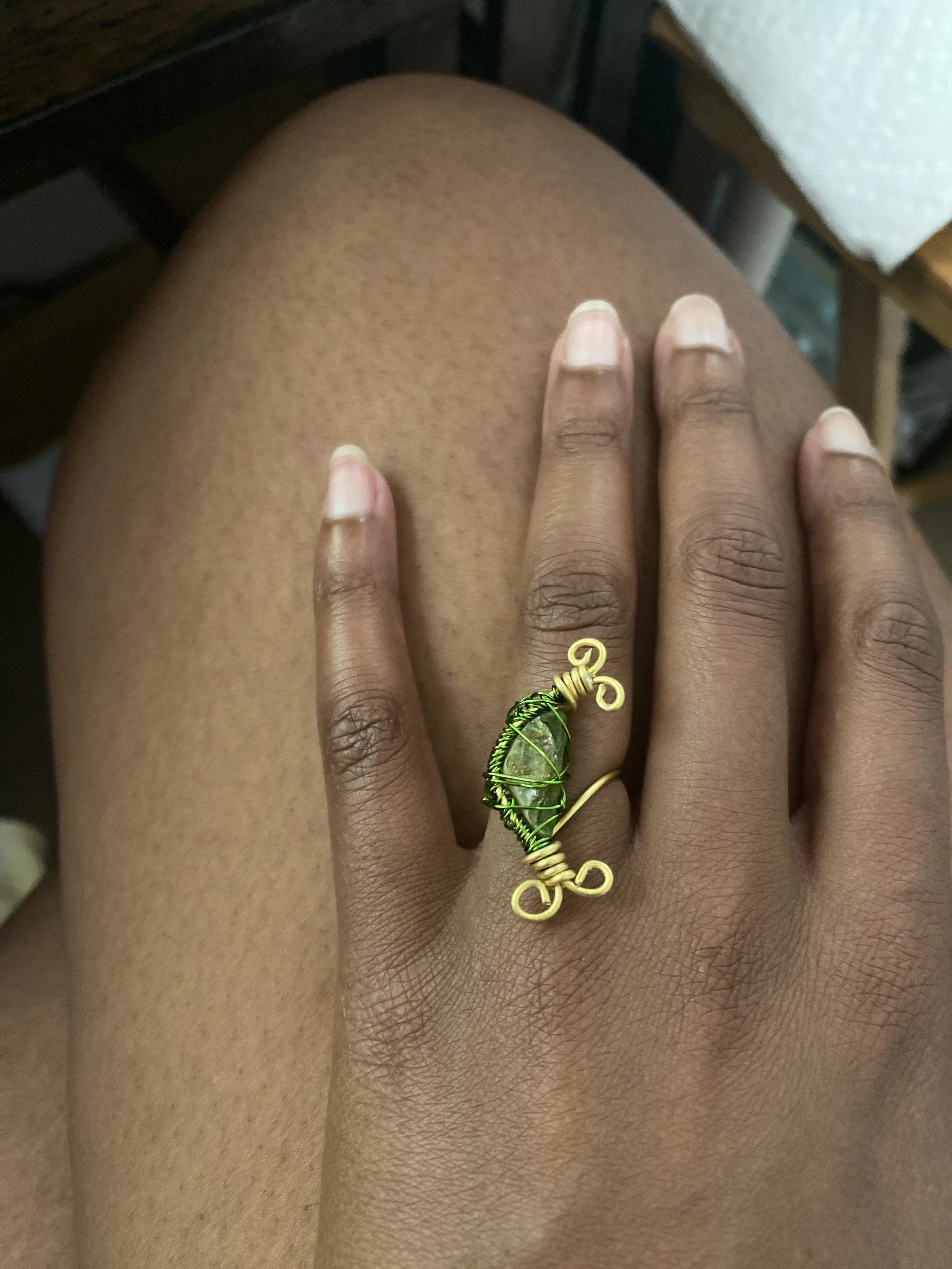 A close-up of a person's hand resting on their knee, wearing a ring with a green stone wrapped in green wire and gold wire embellishments.