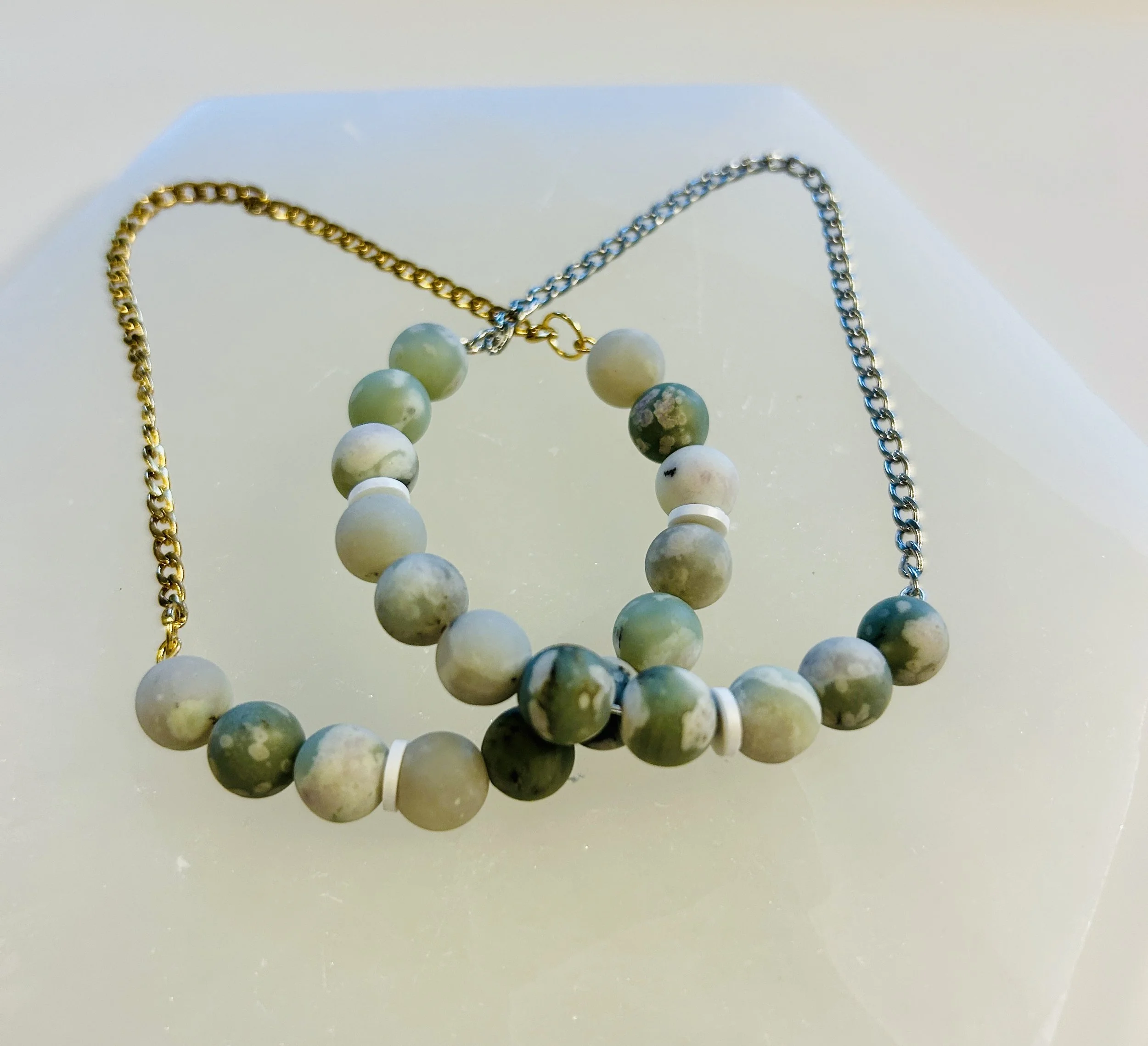 A beaded necklace with green and white marbled beads and a gold and silver chain on a white surface