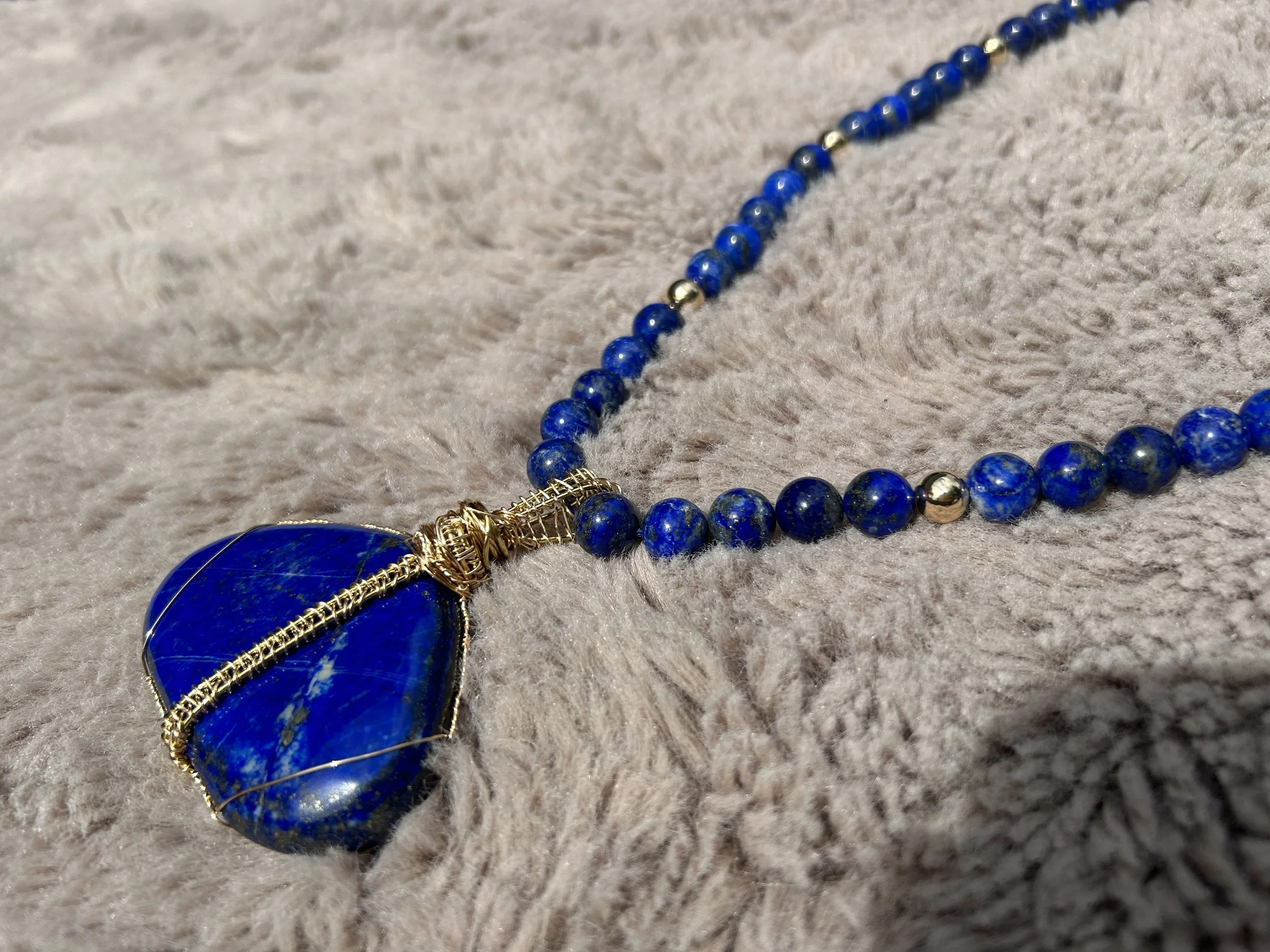 A necklace with a large blue stone pendant wrapped in gold wire, resting on a soft beige fabric background.