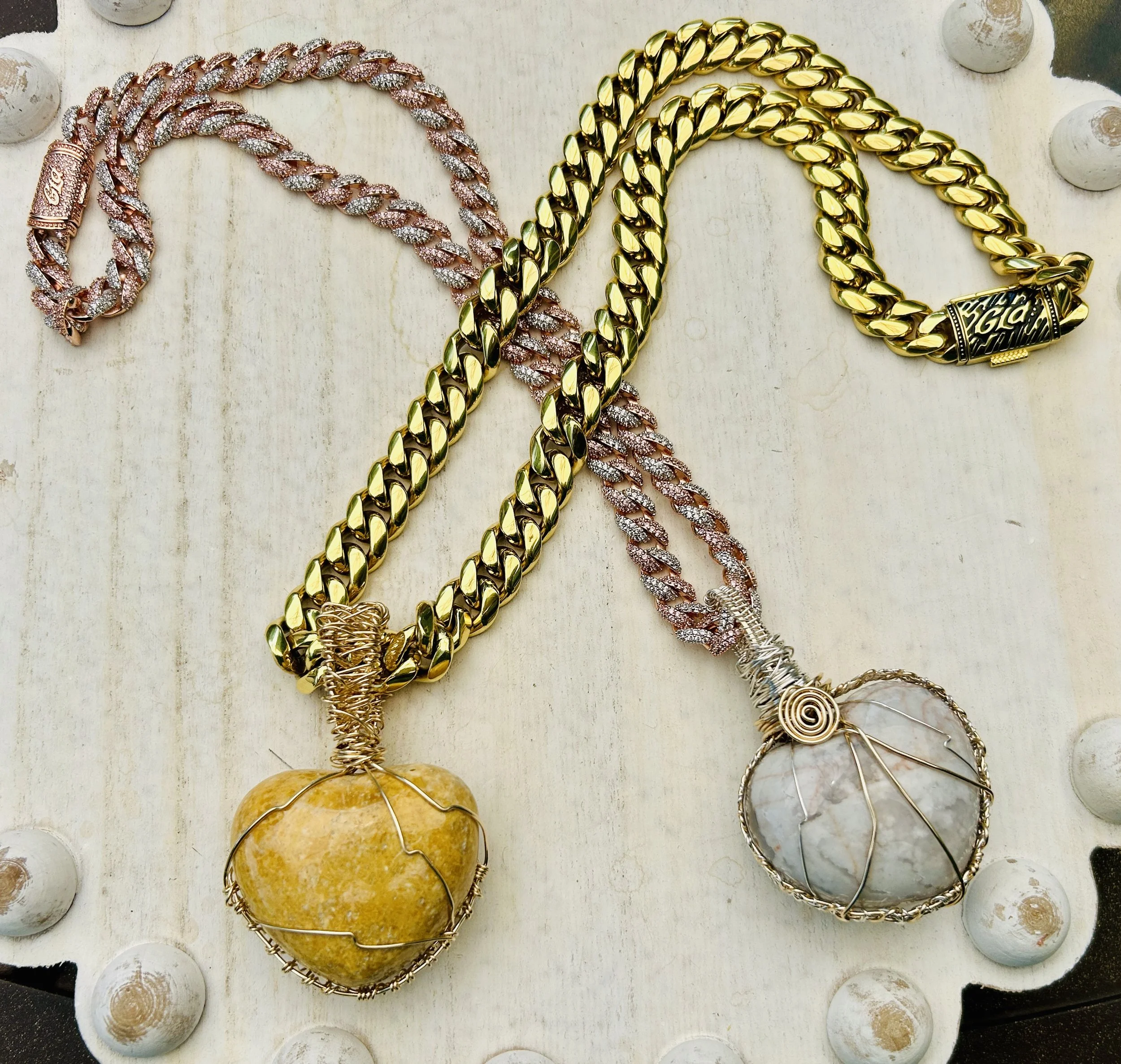 Two chunky gold chains with large pendants, one with a yellow stone and the other with a gray stone, lying on a white surface with scattered round objects.