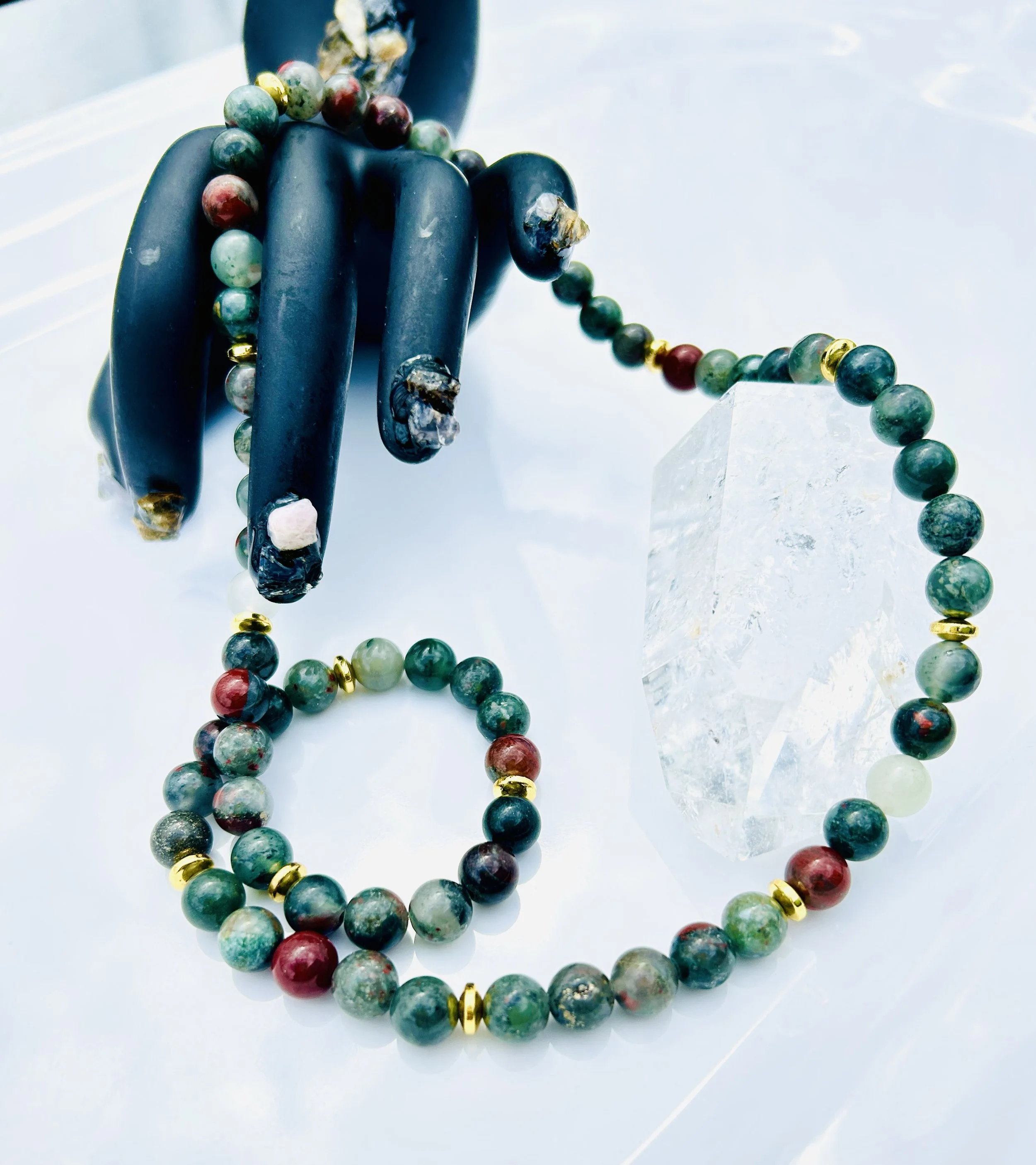 A black hand sculpture holding a beaded necklace and bracelet, with a large clear crystal on a white surface.