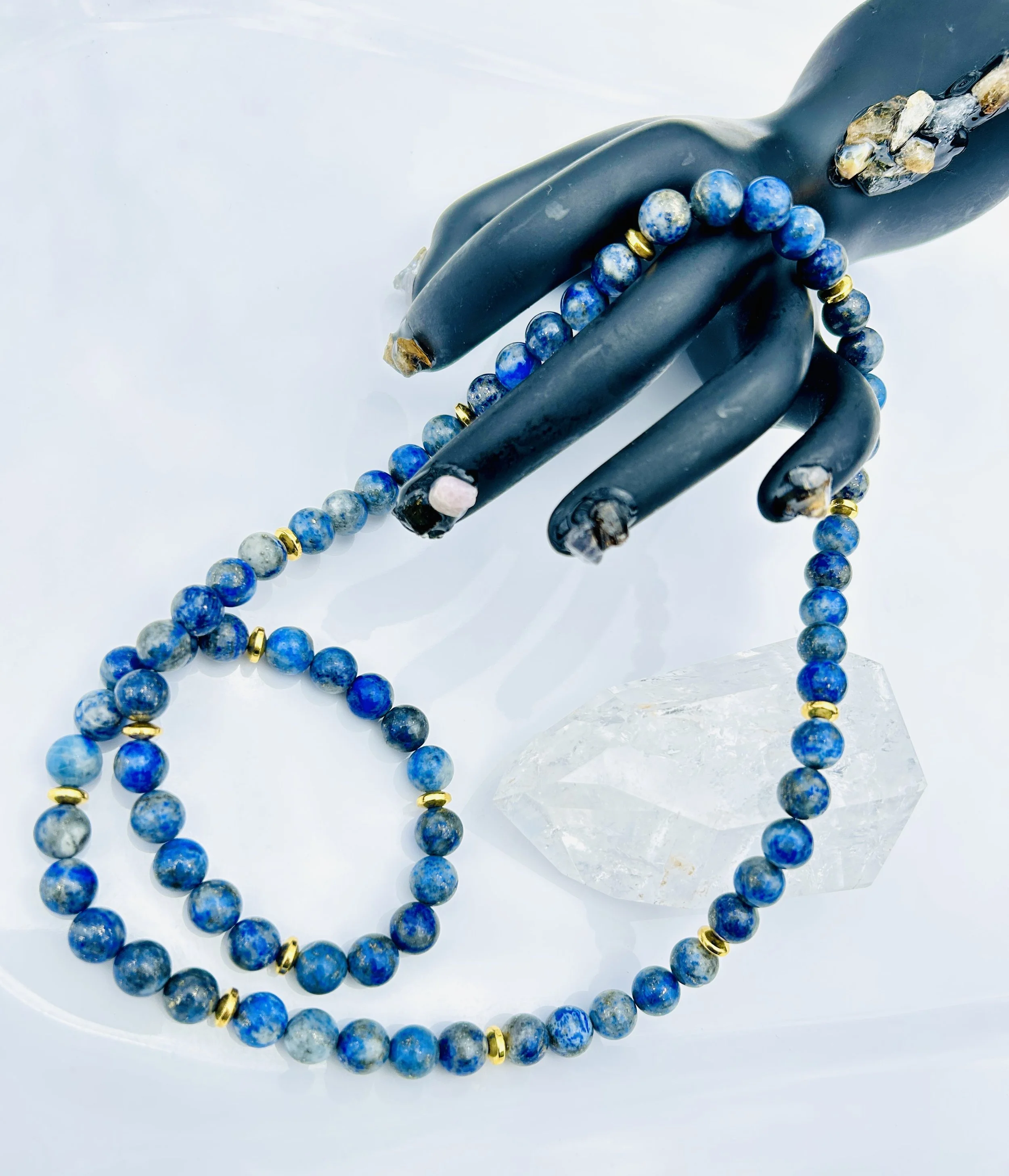 Blue beaded necklace and bracelet with gold accents on black hand-shaped jewelry display, with clear rock crystal on white background.