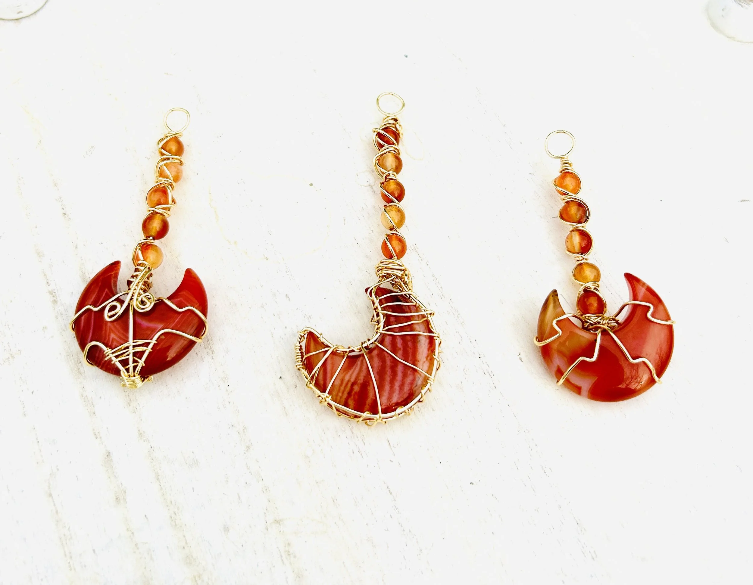Three crescent-shaped jewelry pendants made of red-orange stones wrapped in wire, with beaded loops on top, arranged on a white surface.