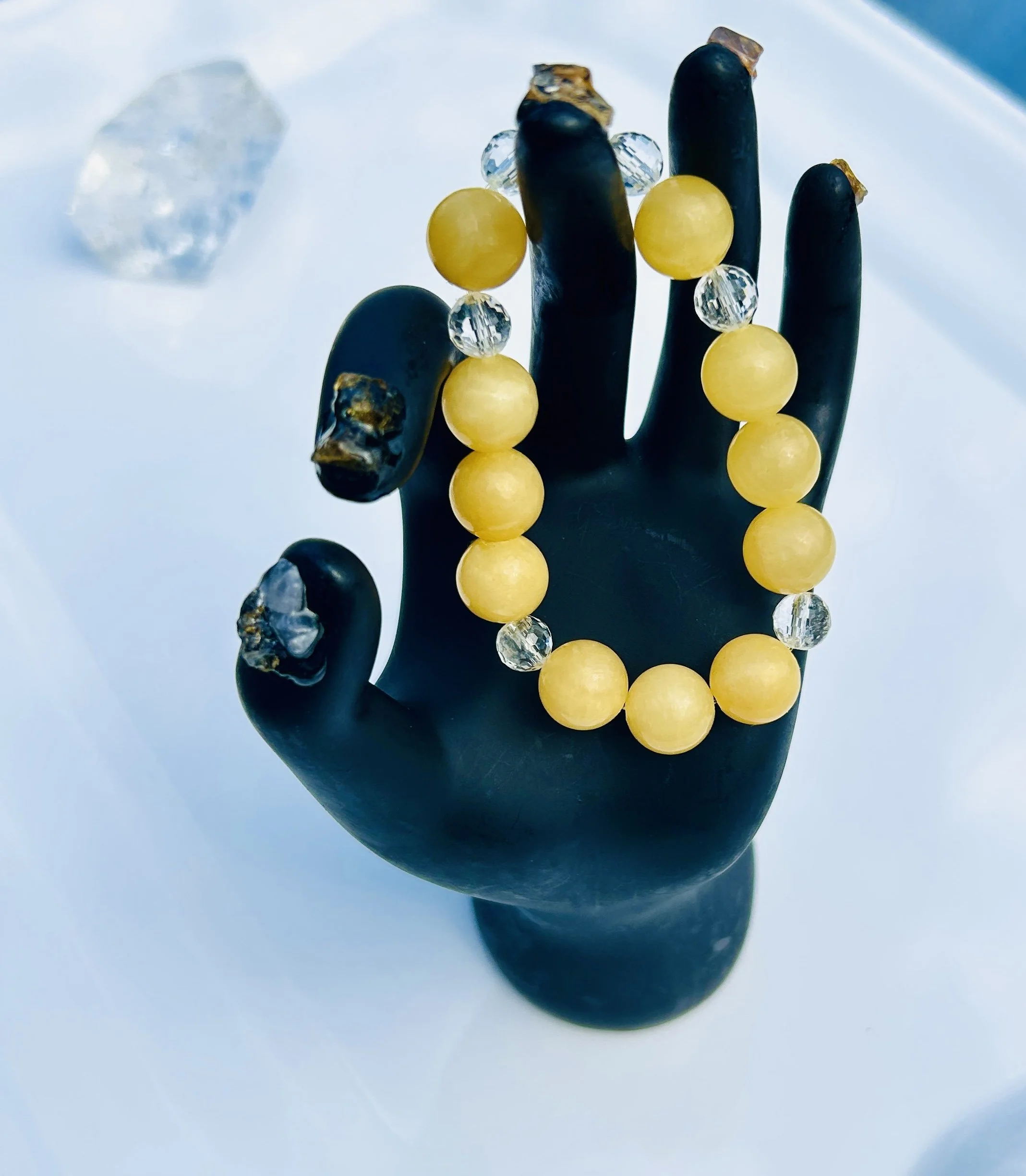 A black mannequin hand displays a beaded bracelet with yellow and clear beads, with some large clear stones in the background.