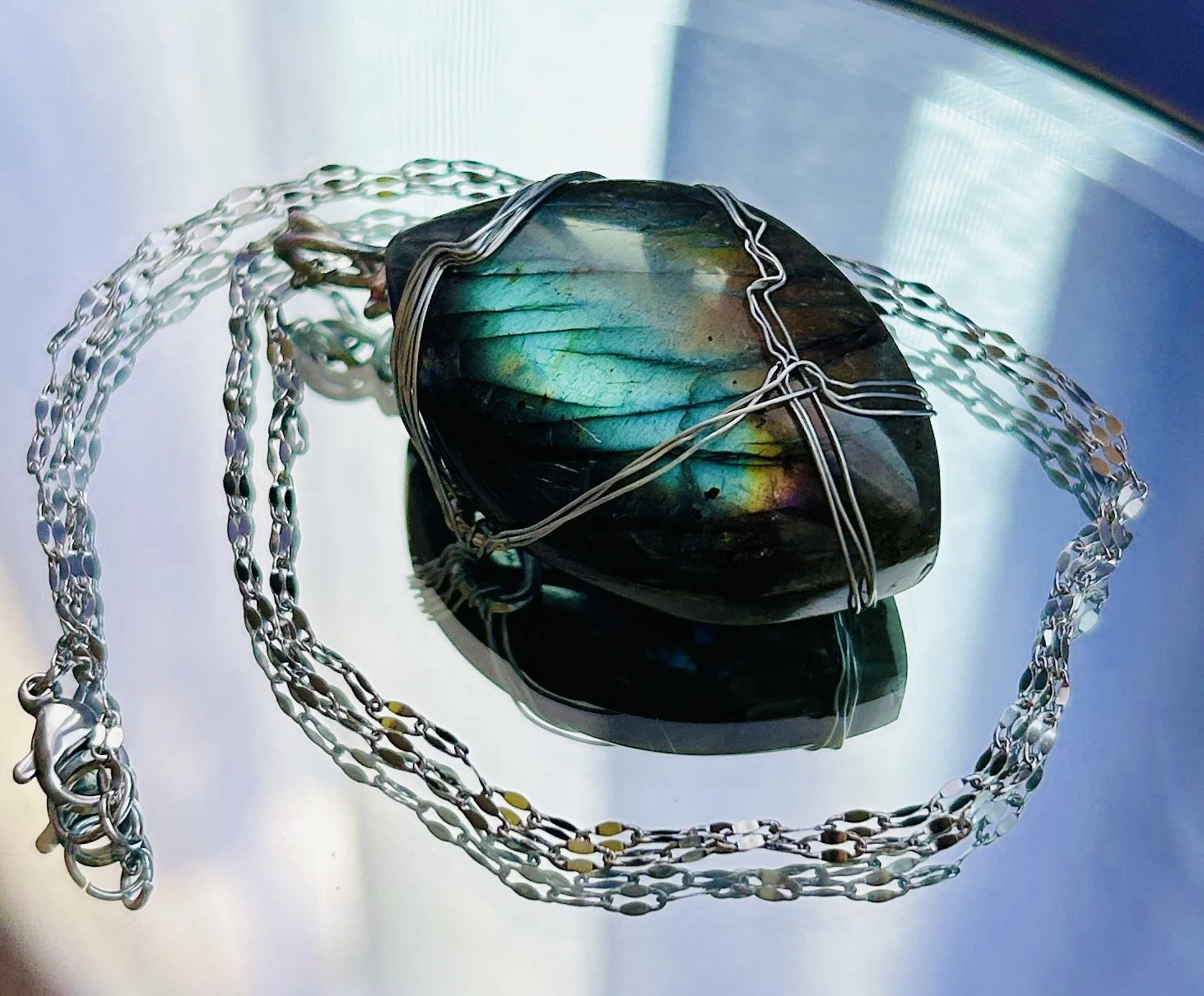 A polished labradorite stone wrapped in wire, with a metallic chain necklace surrounding it, placed on a reflective surface.