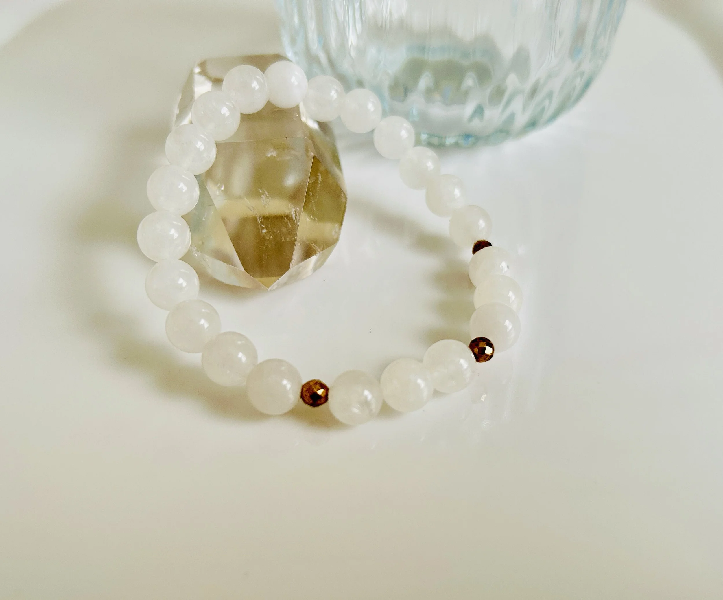 A white beaded bracelet with two small copper-colored beads, a large clear faceted gemstone, and a transparent textured glass container in the background, placed on a white surface.