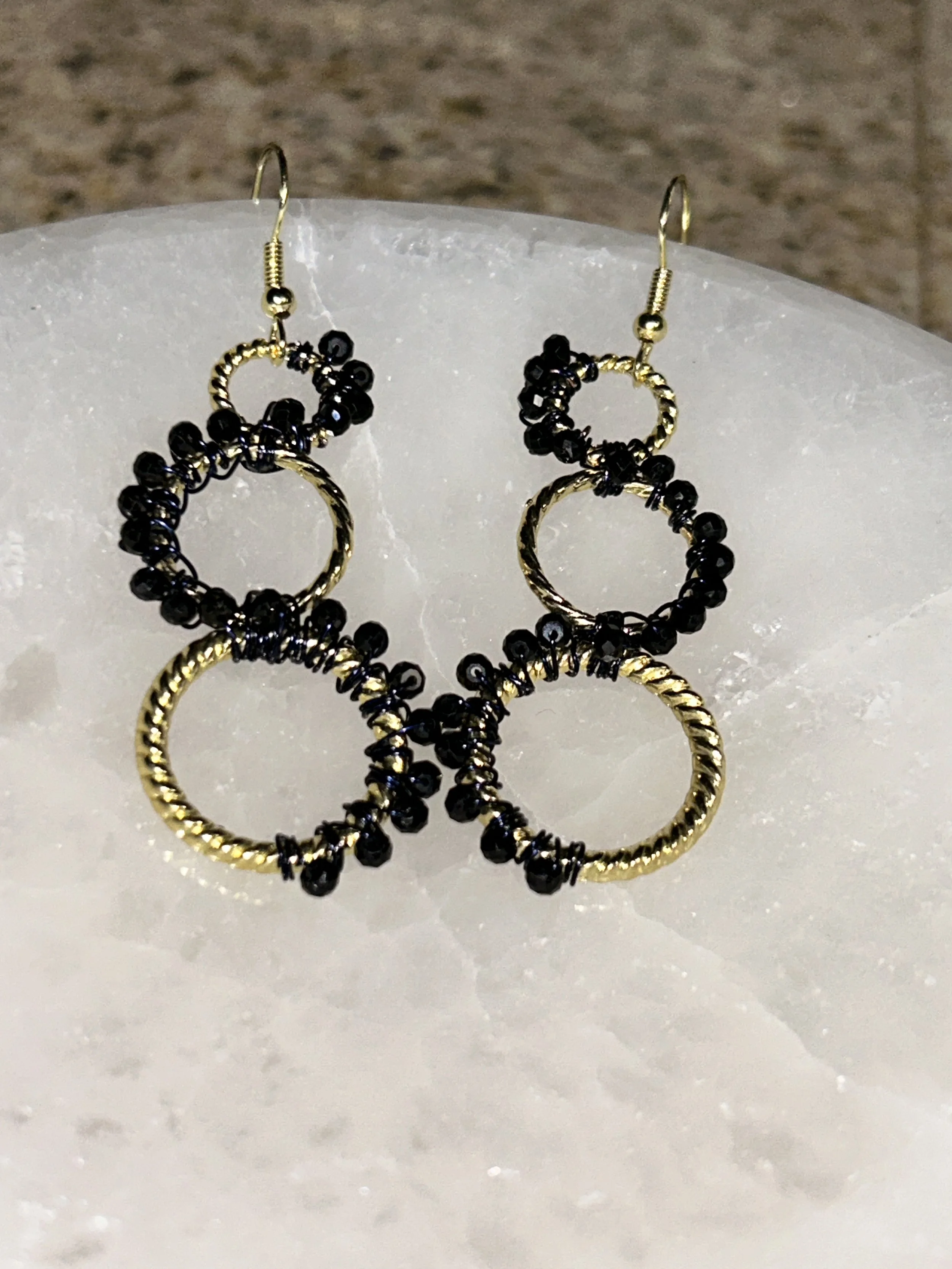 Gold and black hoop earrings with small black beads, displayed on a white stone surface.