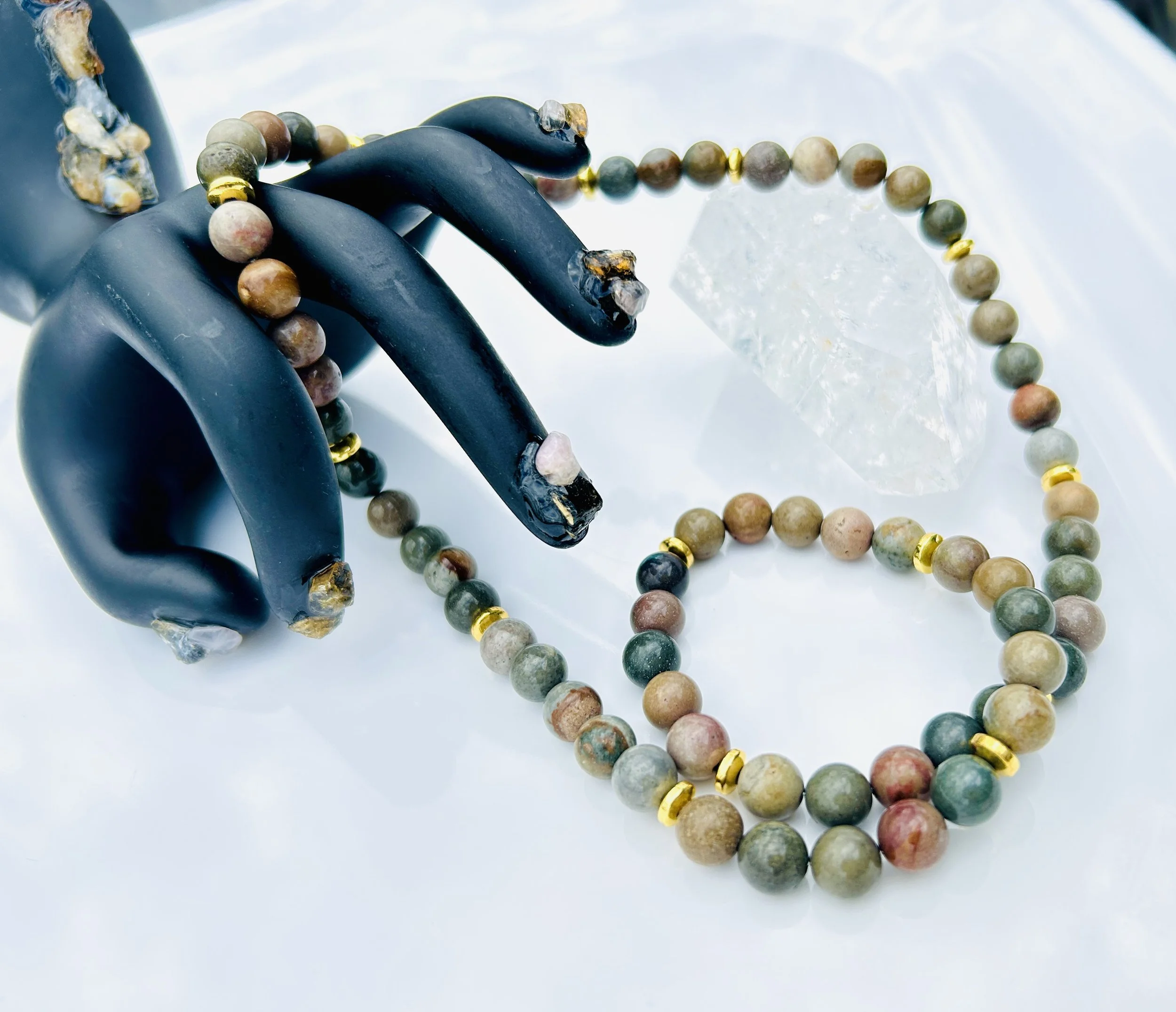 A black snake-shaped object, a beaded necklace with multicolored beads and gold accents, and a clear crystal on a reflective surface.