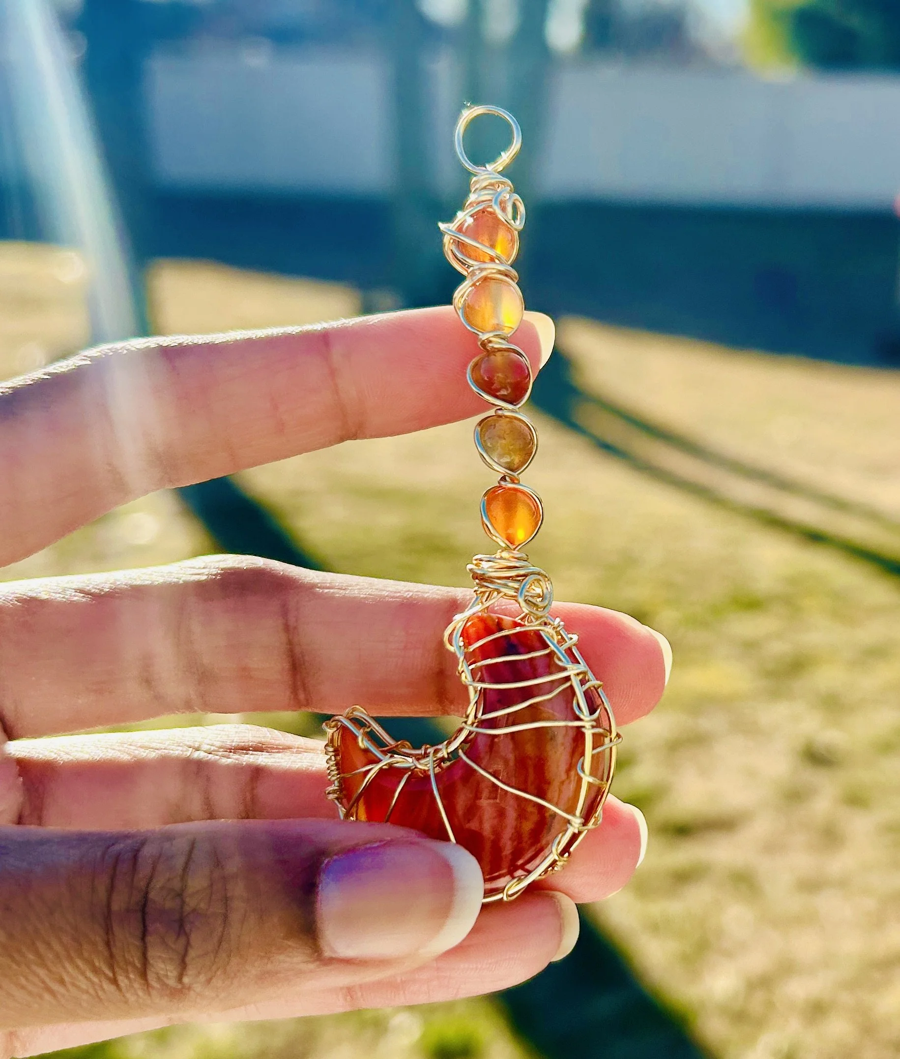 A hand holding a wire-wrapped crescent-shaped pendant with orange stones, set against an outdoor background of a bench and greenery.