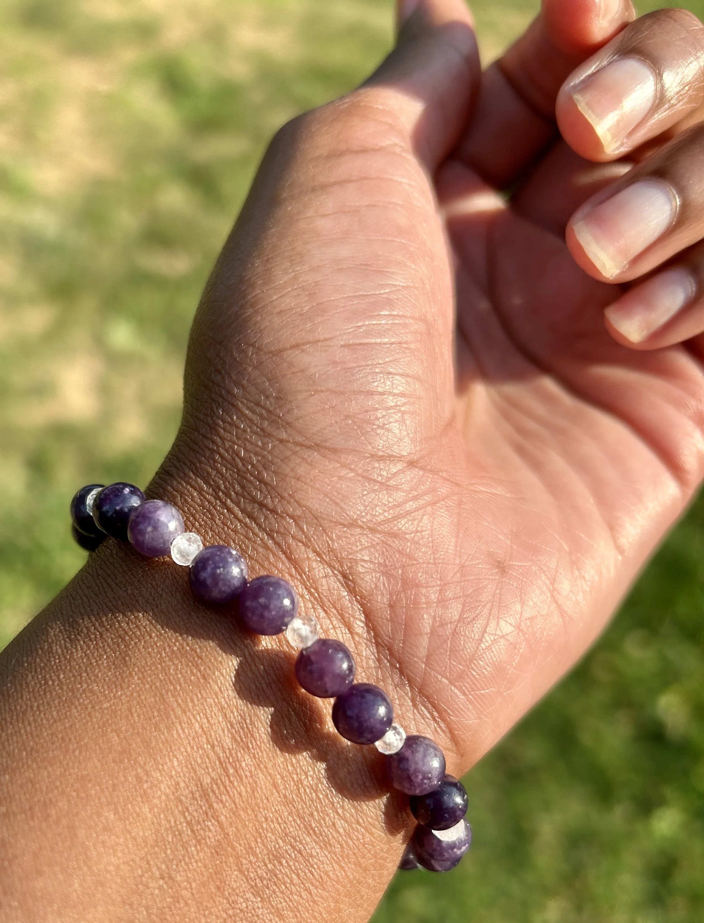 Hand wearing a purple beaded bracelet, outdoors with green grass background.
