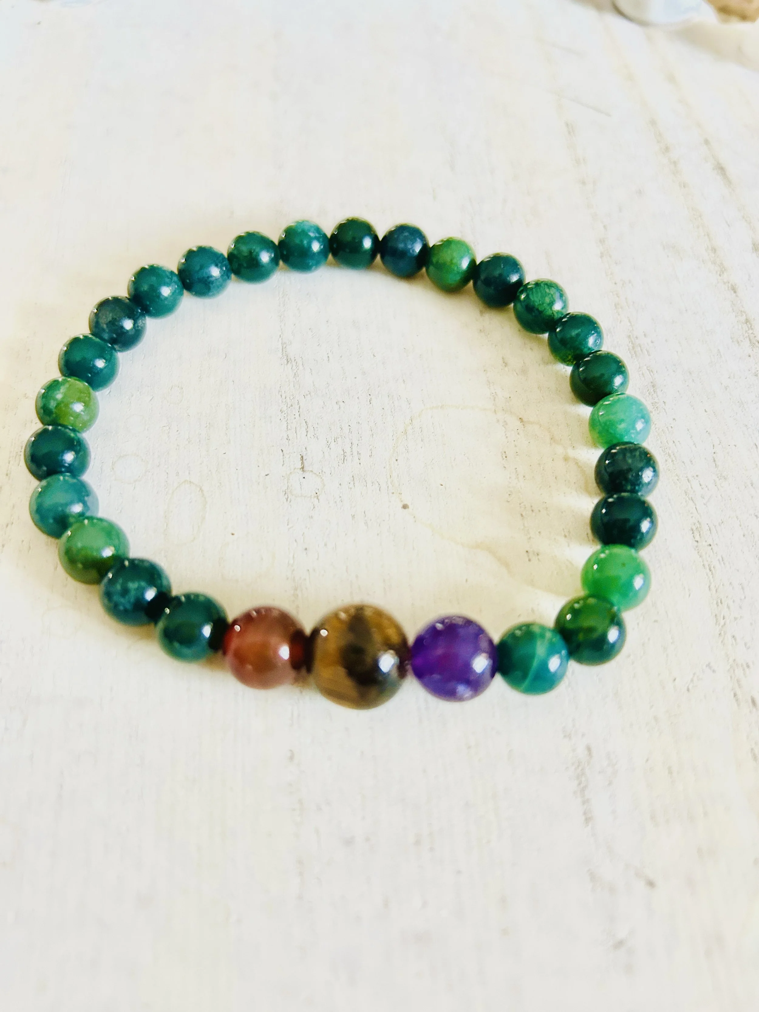 A beaded bracelet with green beads and three larger beads in purple, brown, and red, on a light wooden surface.
