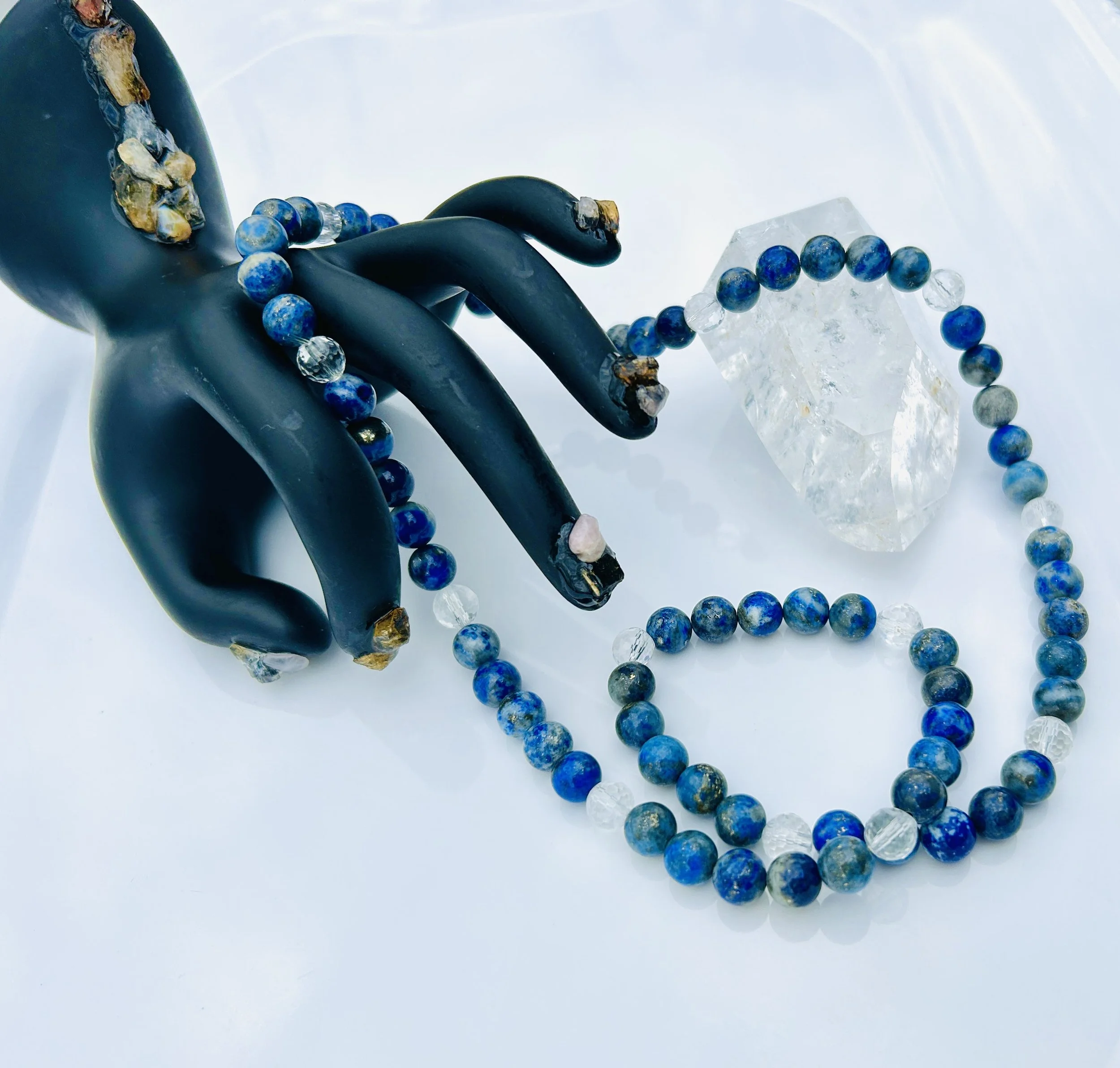 A black hand-shaped jewelry display with a beaded necklace draped over it. The necklace has alternating blue and clear beads and is wrapped around the fingers. There is a large clear quartz crystal positioned beside the display on a white surface.