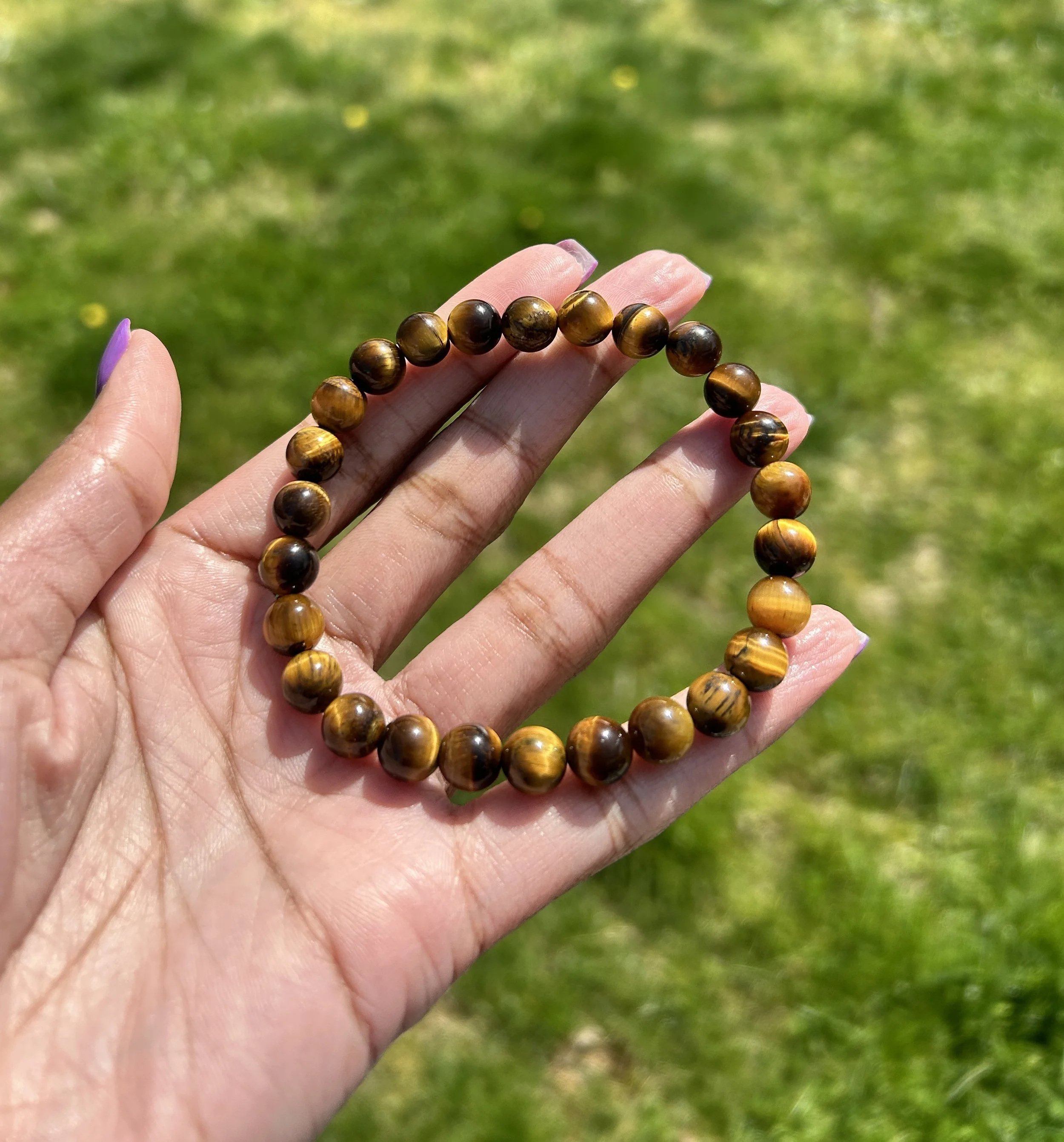 A hand holding a brown and black beaded bracelet outdoors on a sunny day with a grassy background.