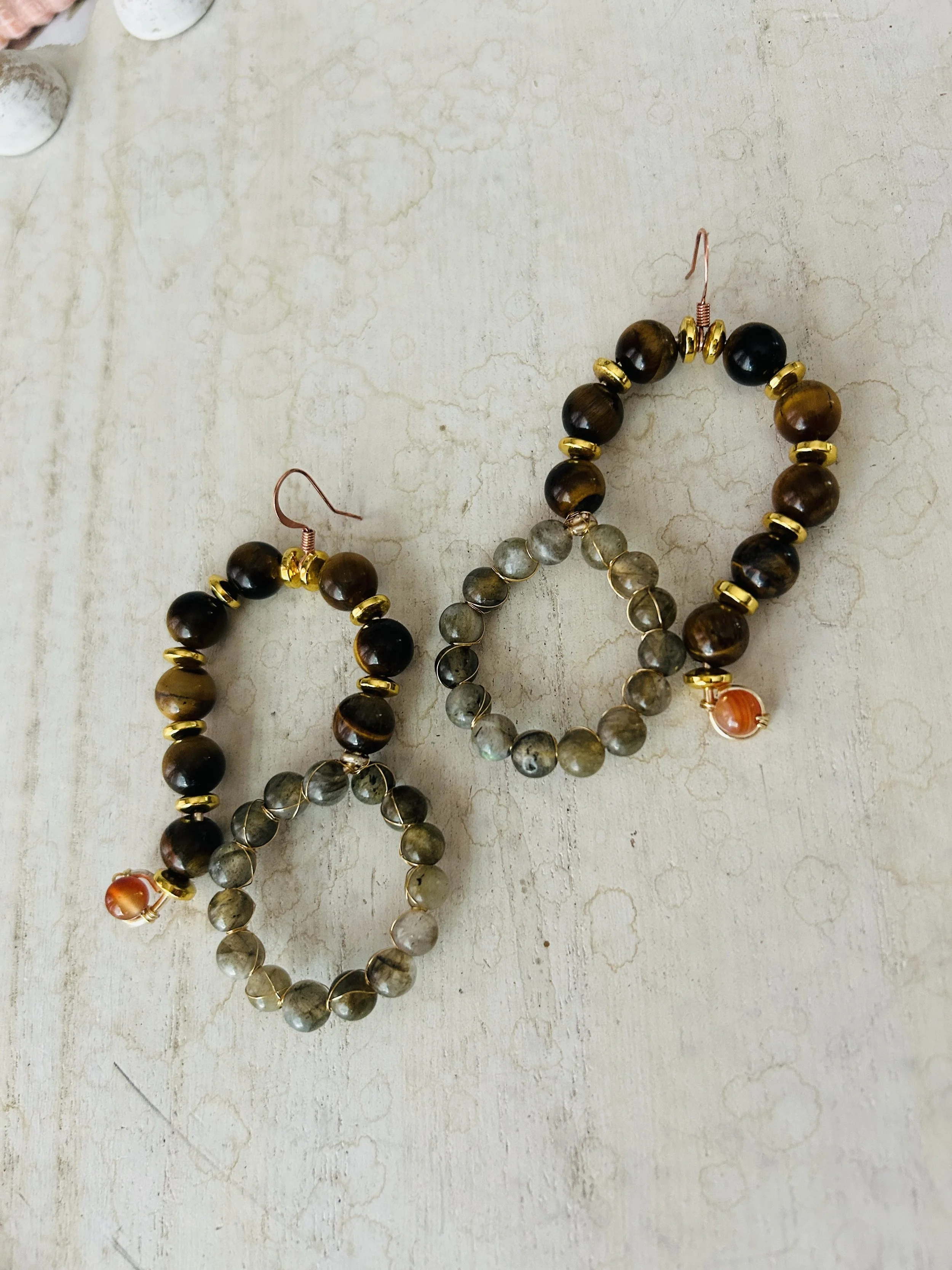 Pair of beaded hoop earrings with dark brown, black, and gray beads, accented with small gold beads and orange beads hanging from hooks, placed on a beige textured surface.
