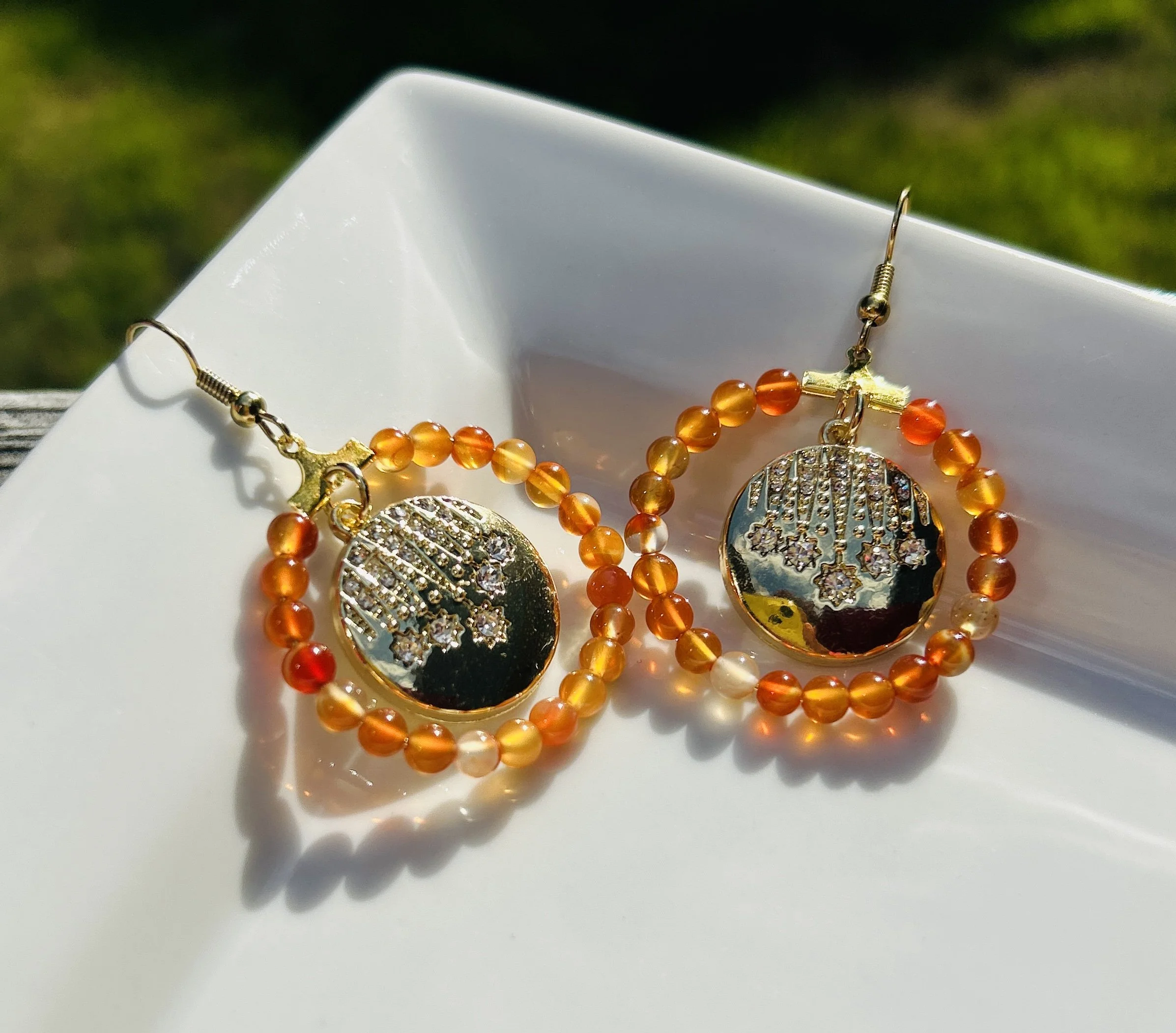 A pair of gold and orange beaded earrings with a circular gold pendant featuring a star and crystal design, displayed on a white dish.