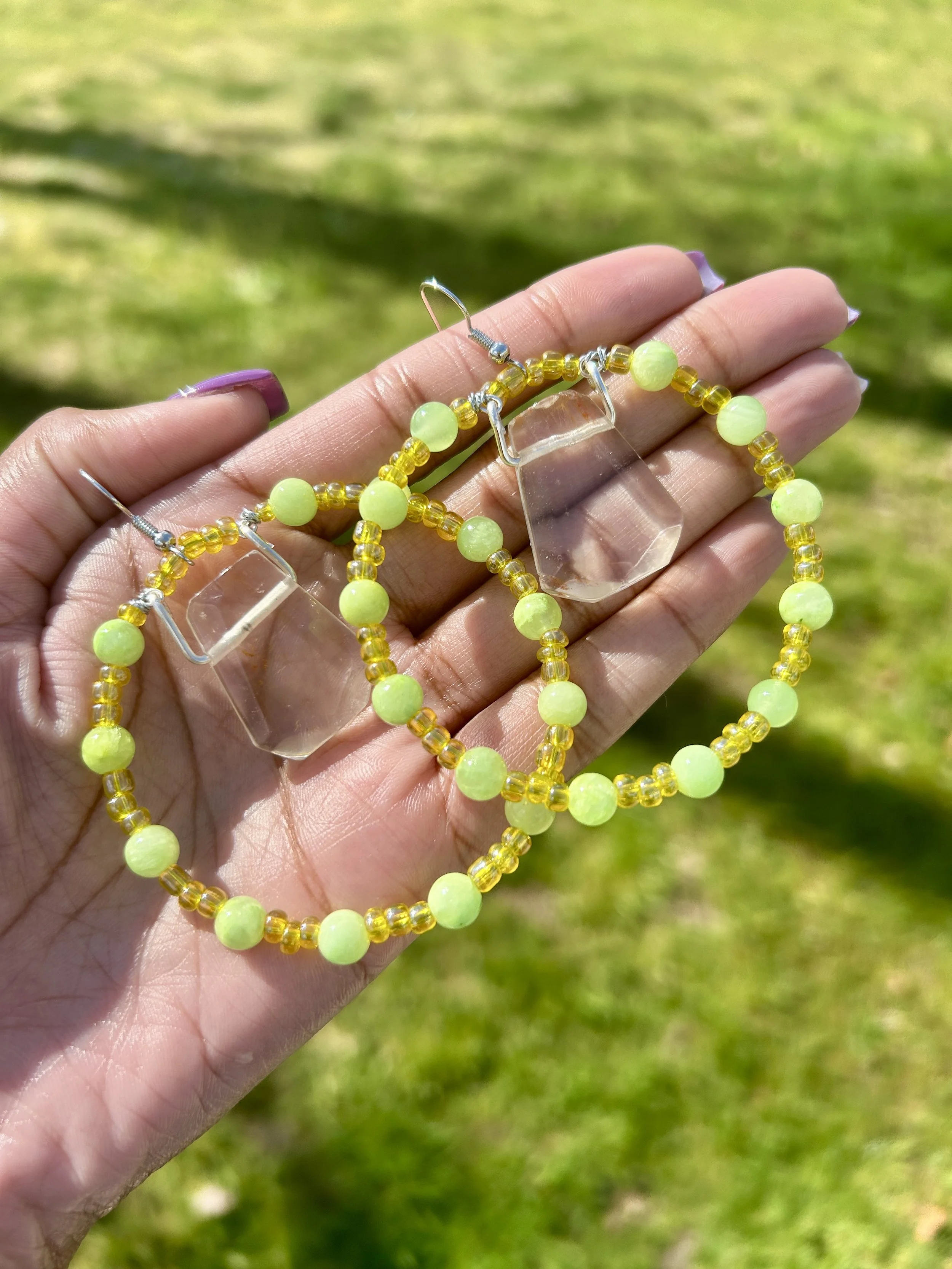 A person's hand holding a pair of yellow and light green beaded hoop earrings with clear plastic hexagonal pendants outdoors on grass.