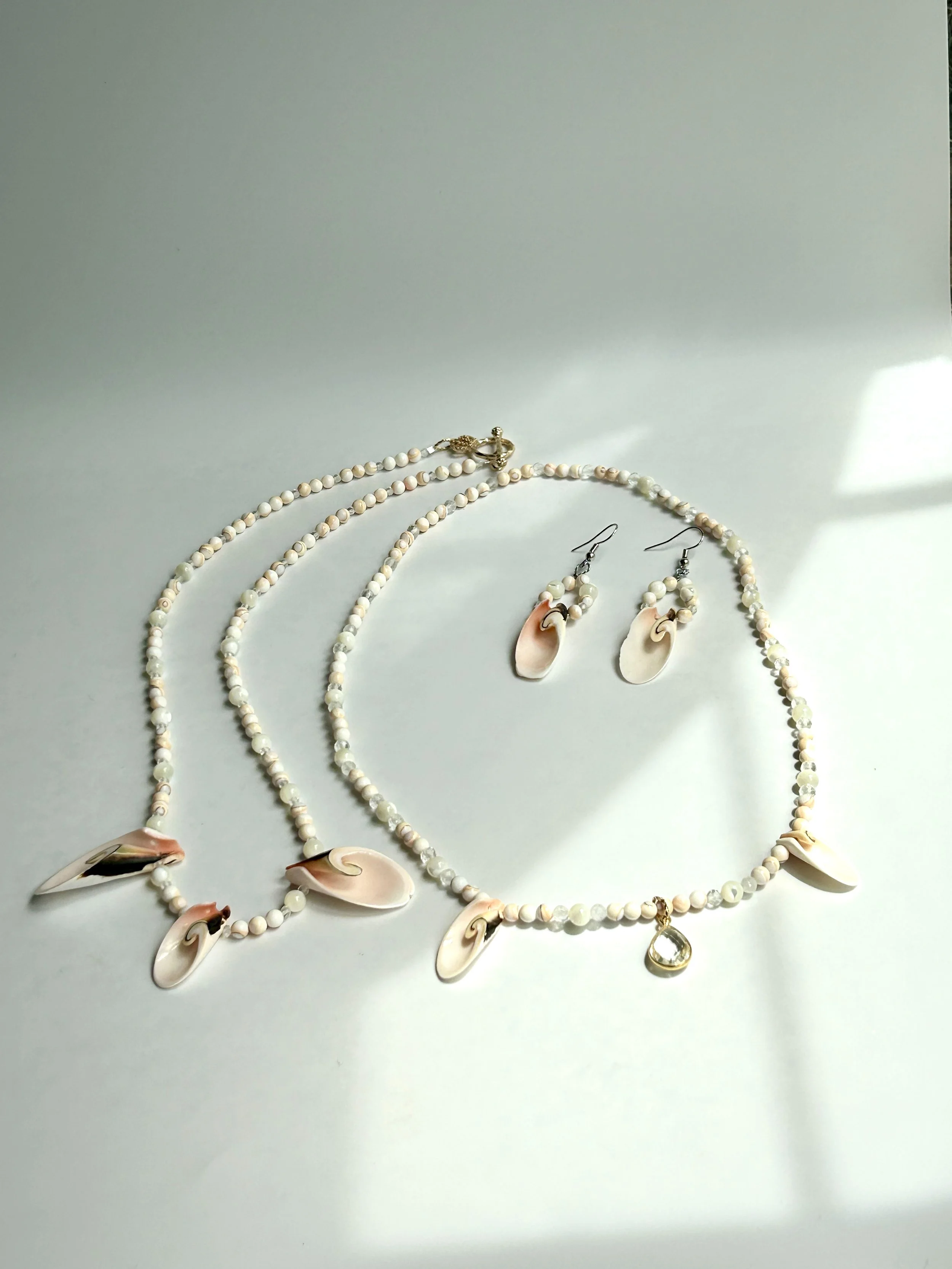 A jewelry set including a necklace, earrings, and a bracelet made of pink and white shells and beads, arranged on a white surface with light and shadow.
